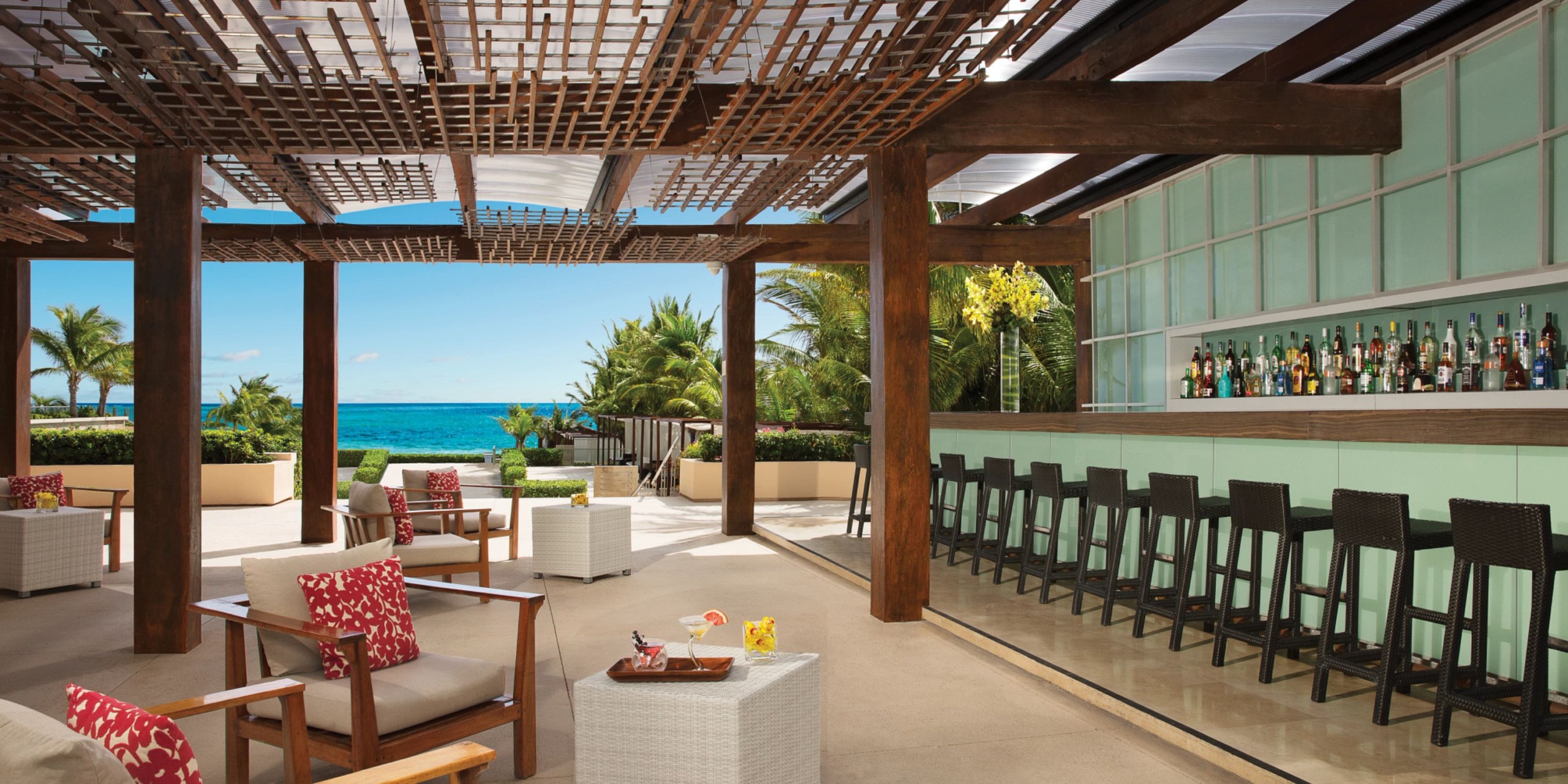 An outdoor bar area at Secrets the Vine Cancun features wooden beams and high stools facing a glass wall, with tropical plants and an ocean view in the background.
