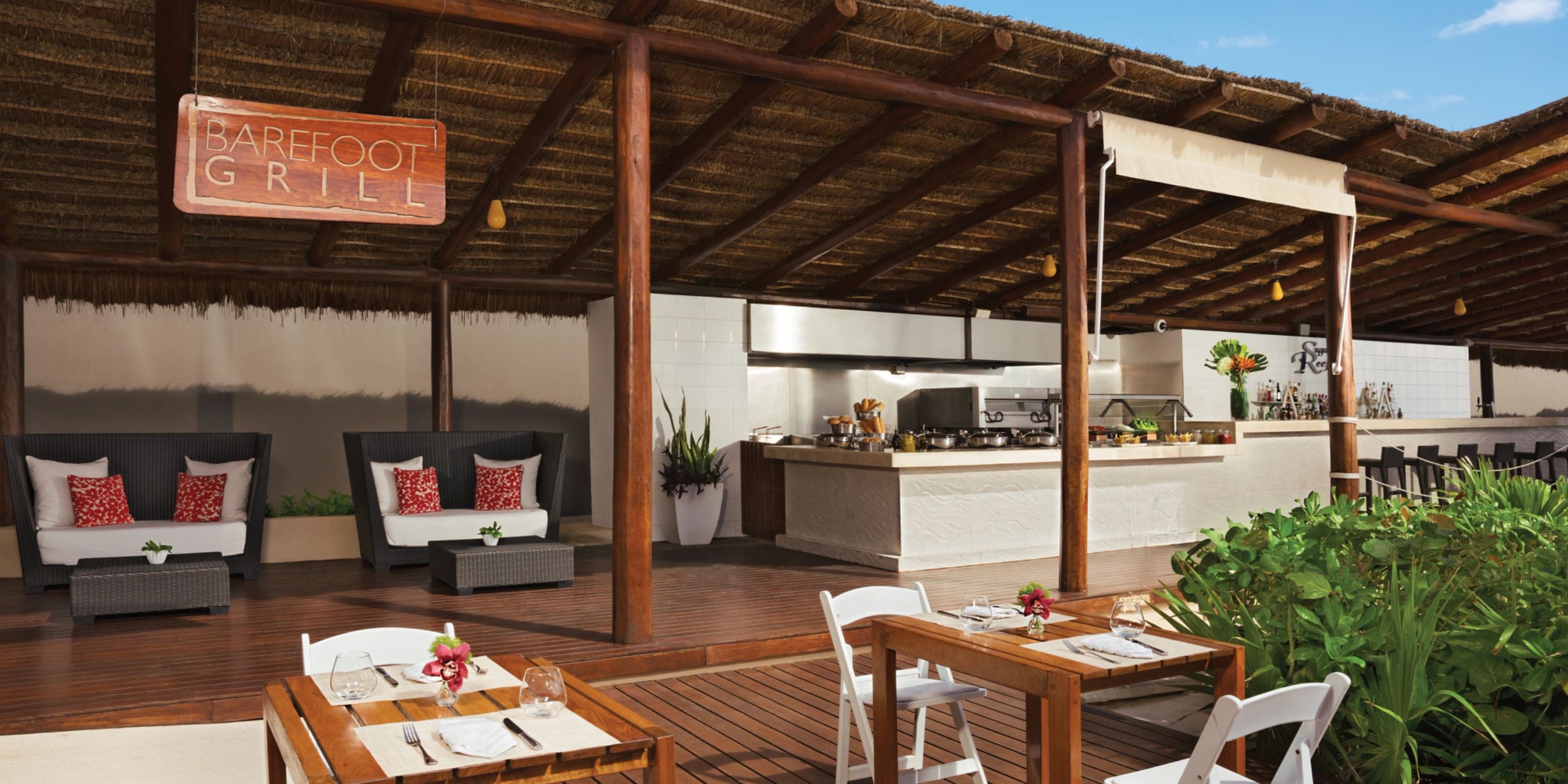 An outdoor restaurant at Secrets the Vine Cancun features a wooden deck, thatched roof, and open kitchen, with tables adorned with flowers. A sign reads "Barefoot Grill.