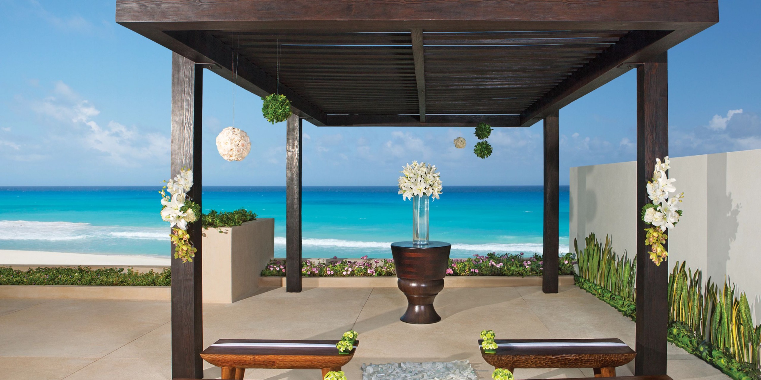 A glimpse of Secrets the Vine Cancun reveals a wooden pergola on a beachfront terrace, adorned with floral arrangements and flanked by benches. A vase of flowers graces the table, all set against the backdrop of the ocean.