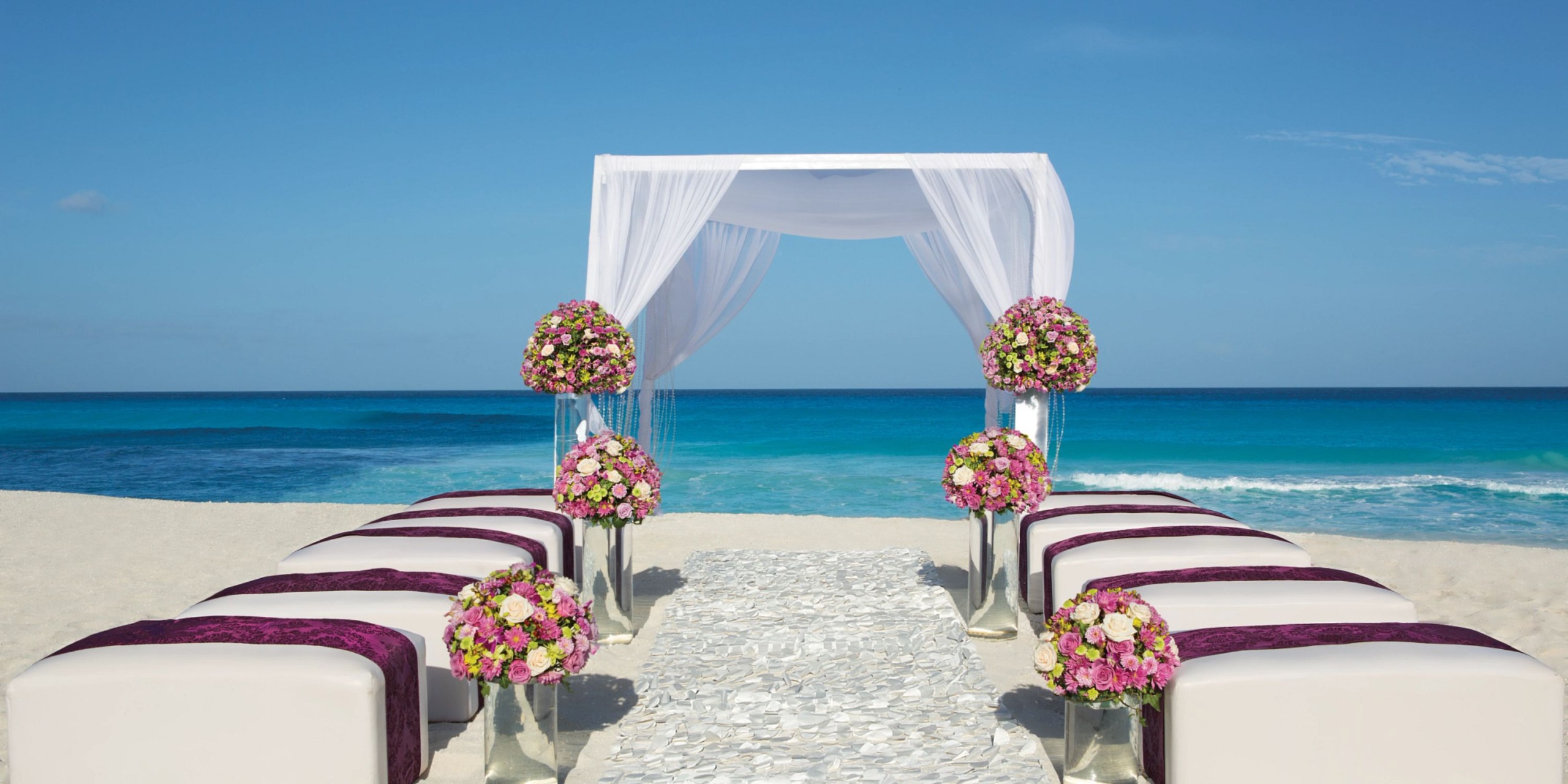 Secrets the Vine Cancun offers an exquisite beach wedding setup featuring a white canopy adorned with floral decorations, overlooking the ocean. Elegant rows of white chairs, each accented with purple sashes, line the aisle leading to a beautifully decorated archway.
