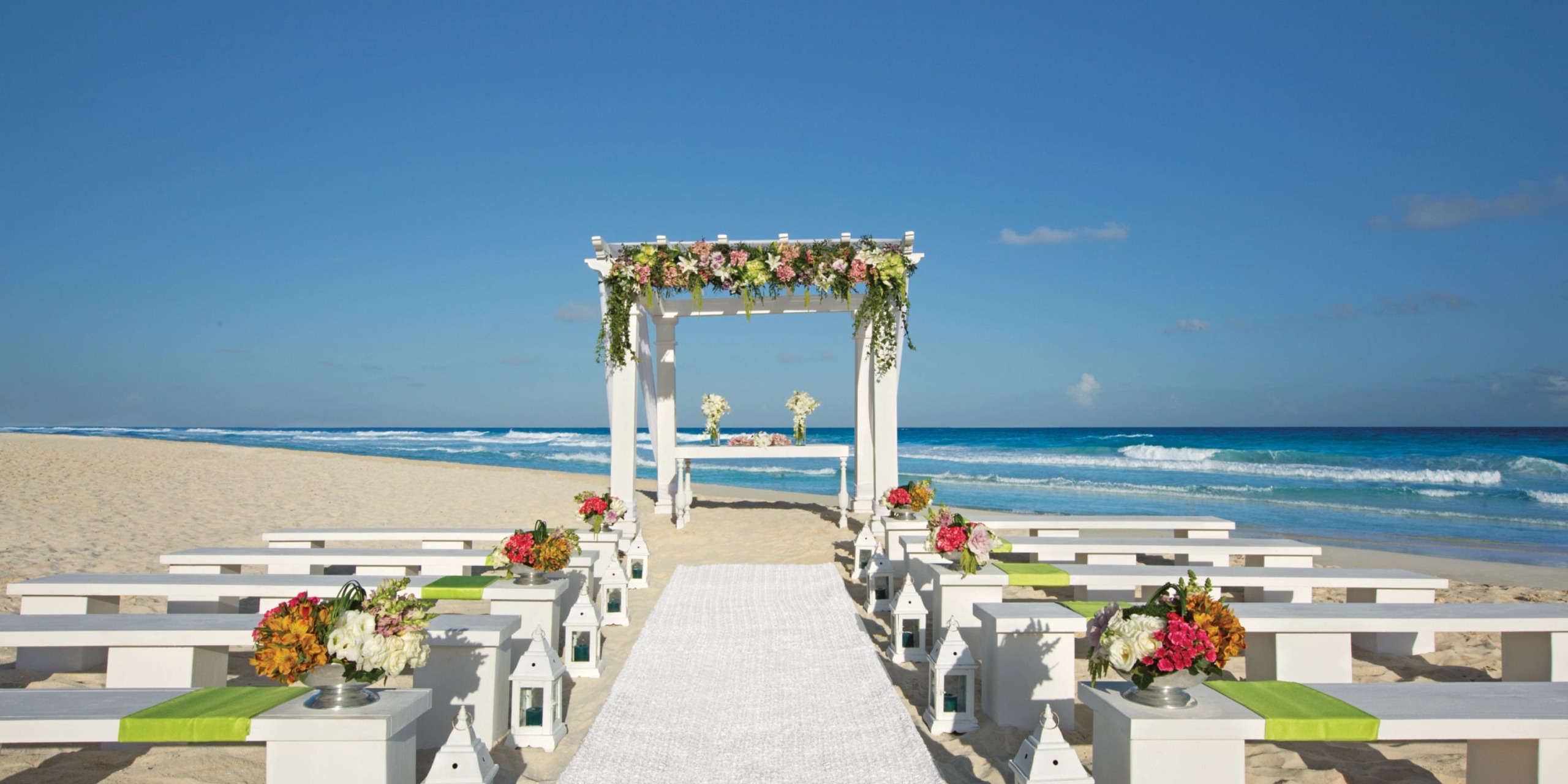 A beach wedding setup at Secrets the Vine Cancun featuring a floral-decorated arch, white benches with green accents, and a white carpet aisle, all set against the stunning backdrop of blue sky and ocean waves.