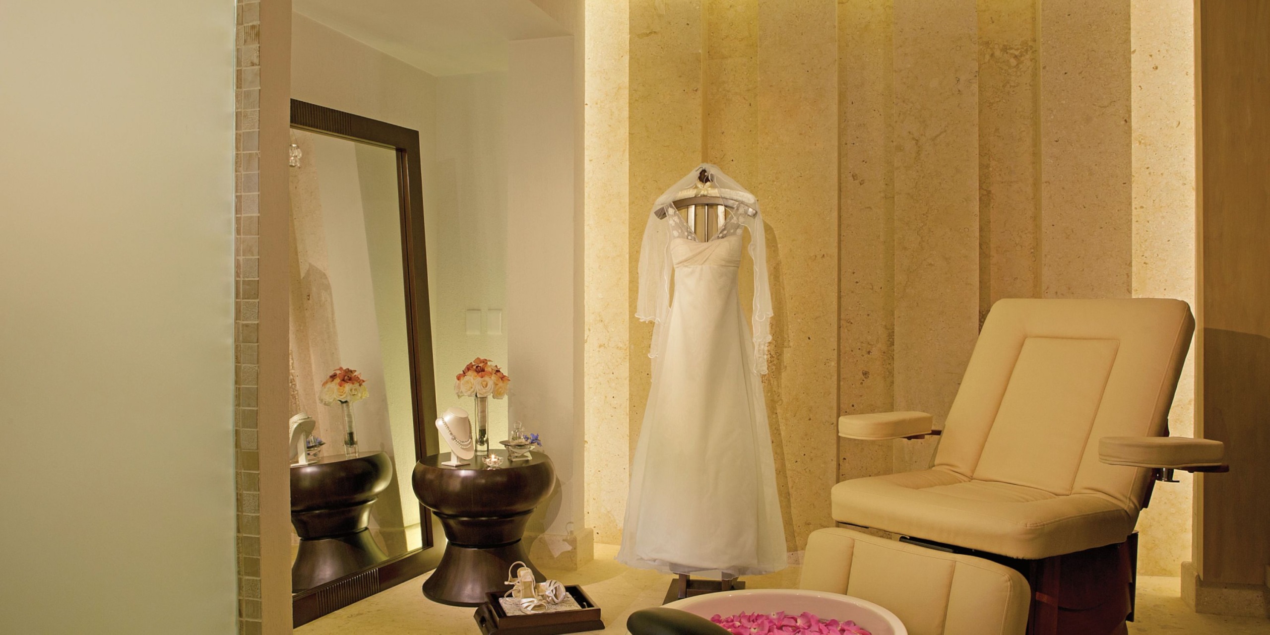 A Secrets the Vine Cancun bridal gown on a stand beside a large mirror, adorned with flower arrangements on small tables and paired with a cream-colored chair in a softly lit room.