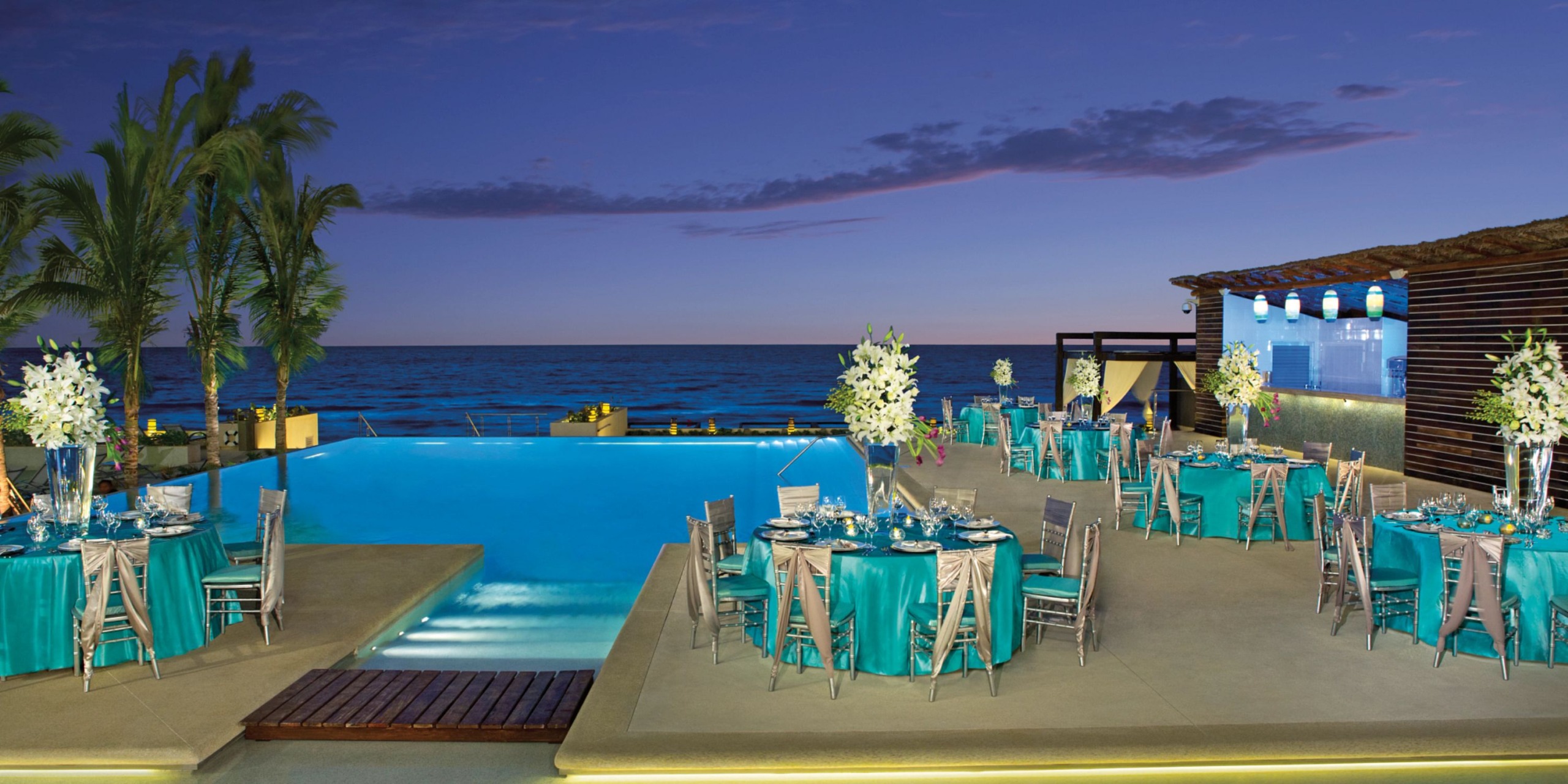 Set up your outdoor event at Secrets the Vine Cancun by an infinity pool, featuring round tables adorned with teal tablecloths and elegant white floral centerpieces, all with a stunning ocean view backdrop under a dusky sky.
