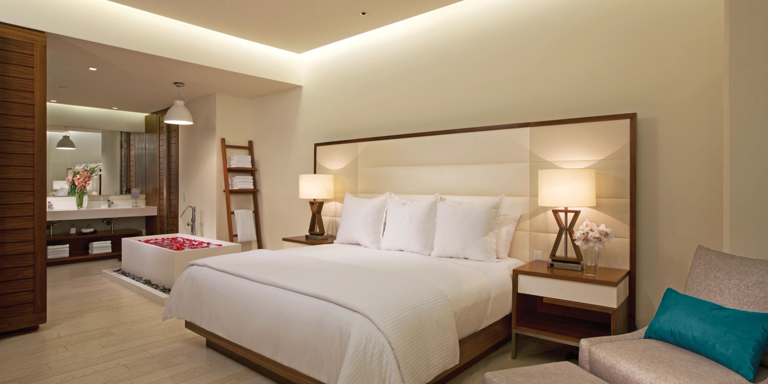 Experience a contemporary room at Secrets the Vine Cancun, featuring a king-sized bed dressed in crisp white linens and conveniently placed bedside lamps. In the background, a bathroom complete with a tub and an elegant ladder shelf is also visible.