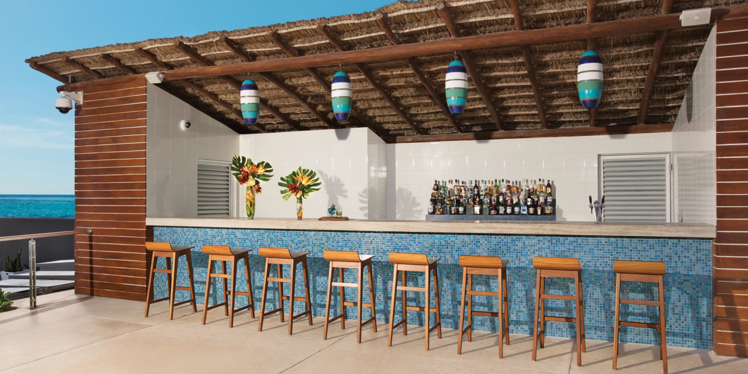 The outdoor beach bar at Secrets the Vine Cancun features a thatched roof, wooden stools, and a tiled counter. Behind the bar, bottles are neatly arranged, while hanging lights illuminate the space. The ocean provides a stunning backdrop in the distance.