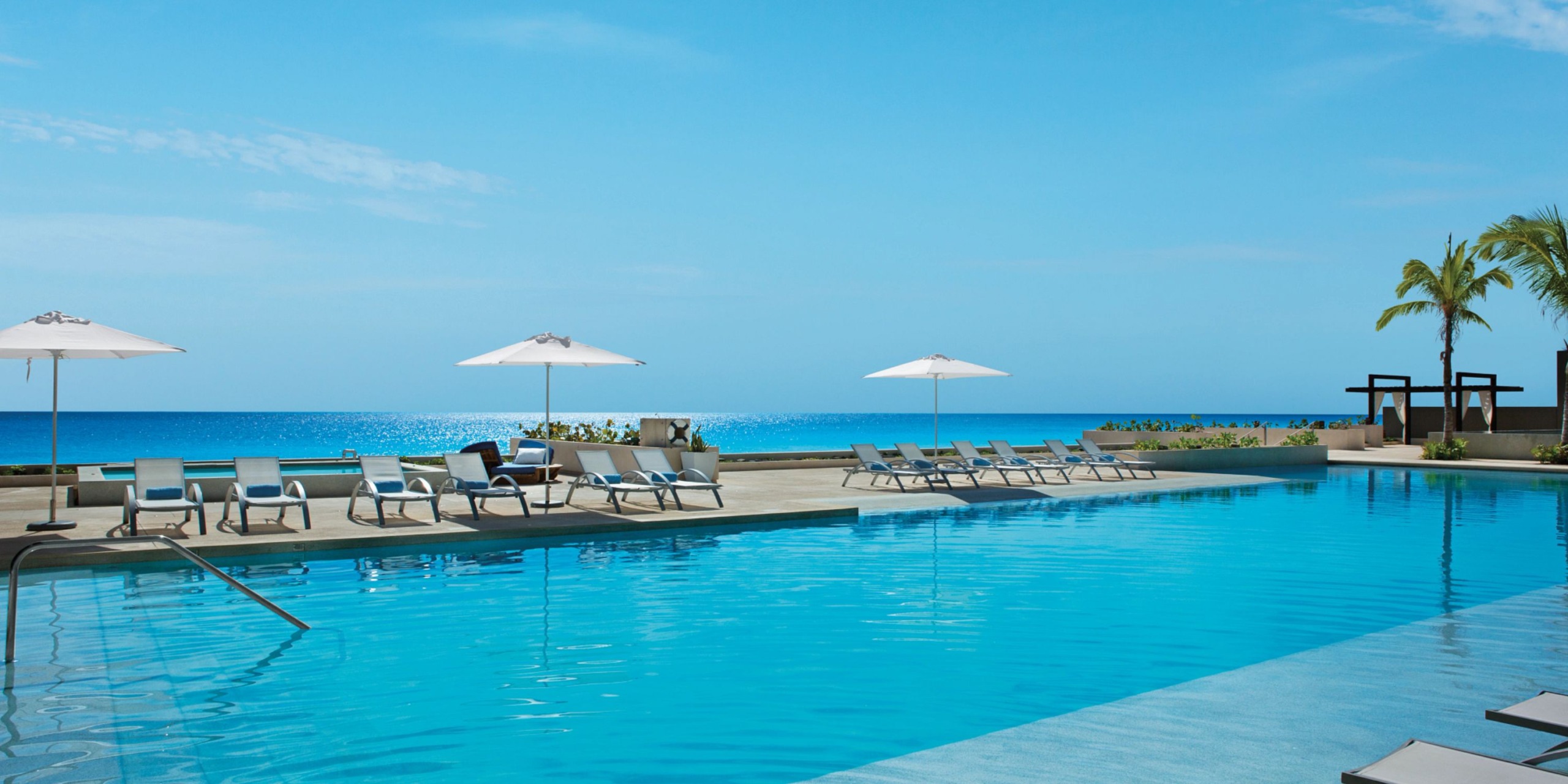 Experience tranquility at Secrets the Vine Cancun, where an outdoor pool with lounge chairs and umbrellas offers stunning views of the calm ocean under a clear blue sky.