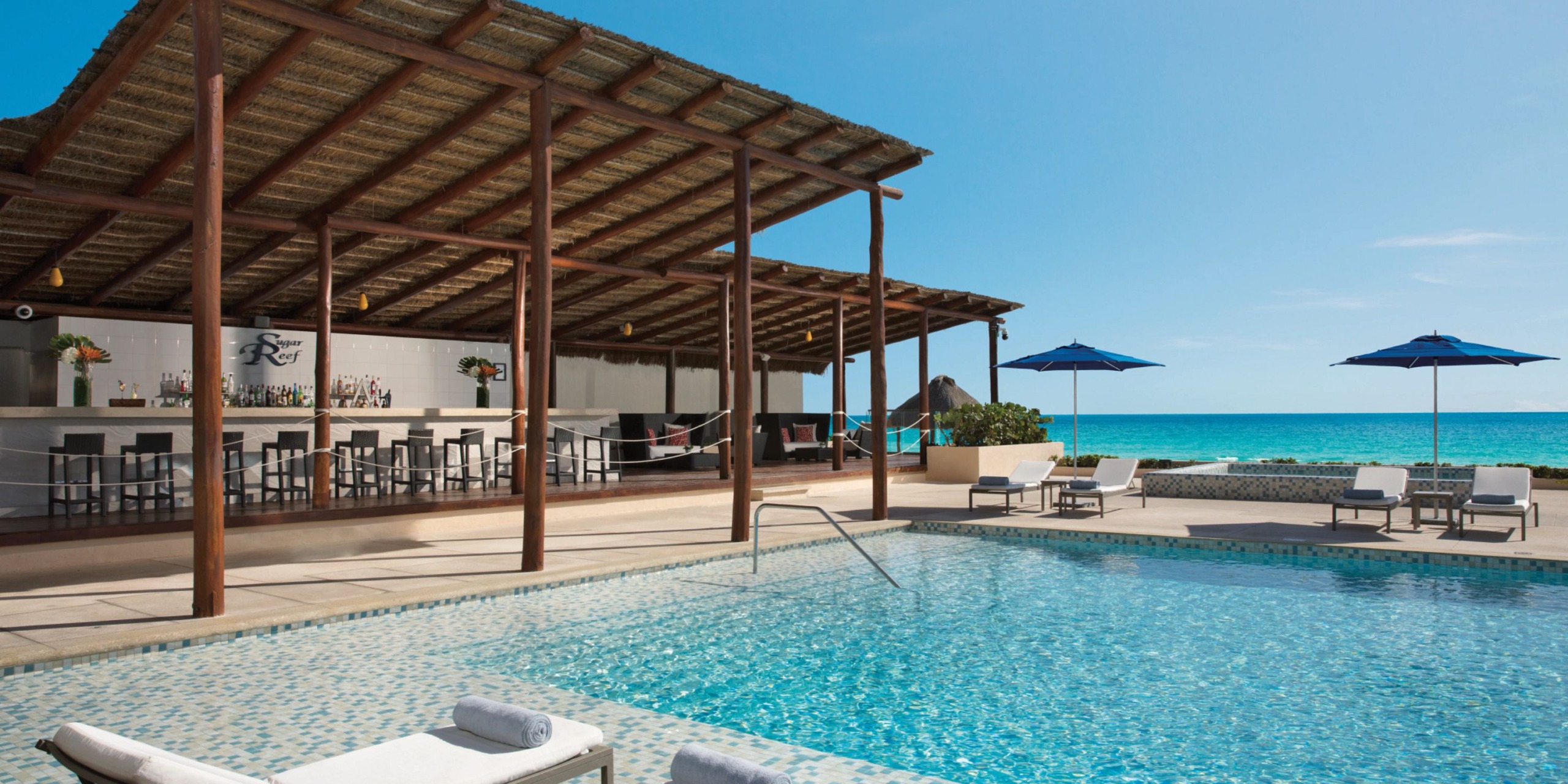 Secrets the Vine Cancun's outdoor pool area features lounge chairs, umbrellas, and a thatched-roof bar with stunning ocean views on a clear day.