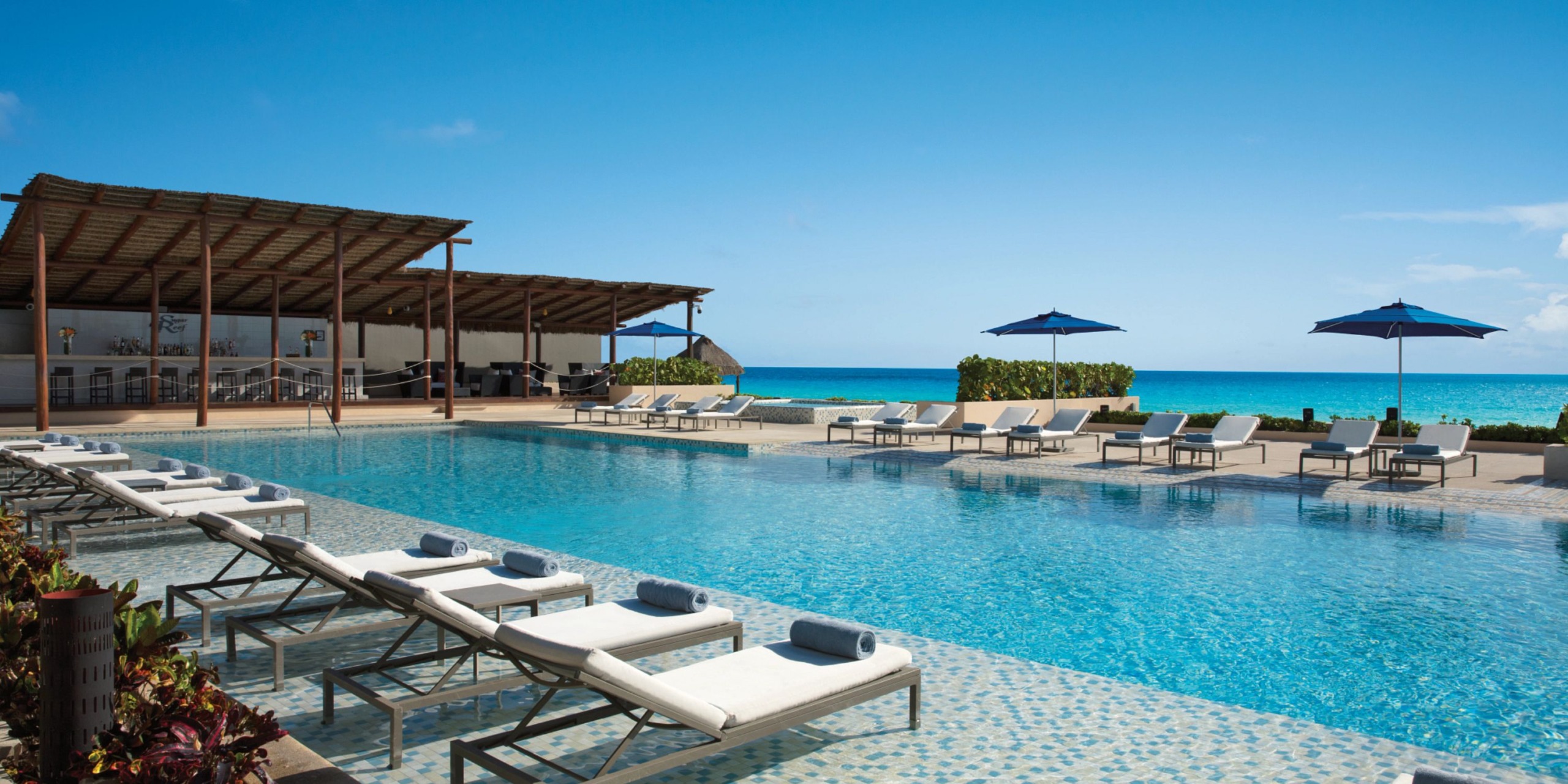 Discover the outdoor pool area at Secrets the Vine Cancun, featuring lounge chairs and umbrellas by the ocean. To the left, find a covered seating area perfect for relaxation. Enjoy the clear sky and serene waters.