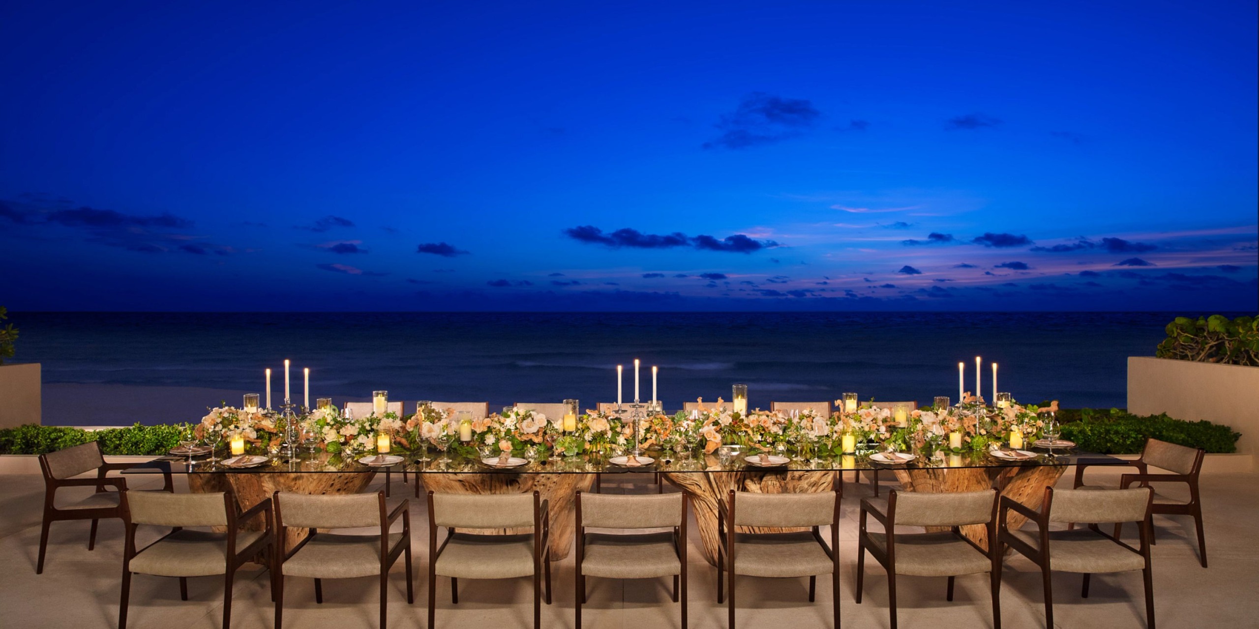 A long, elegantly set table with candles and floral arrangements is positioned outdoors at Secrets the Vine Cancun, overlooking the ocean at dusk.