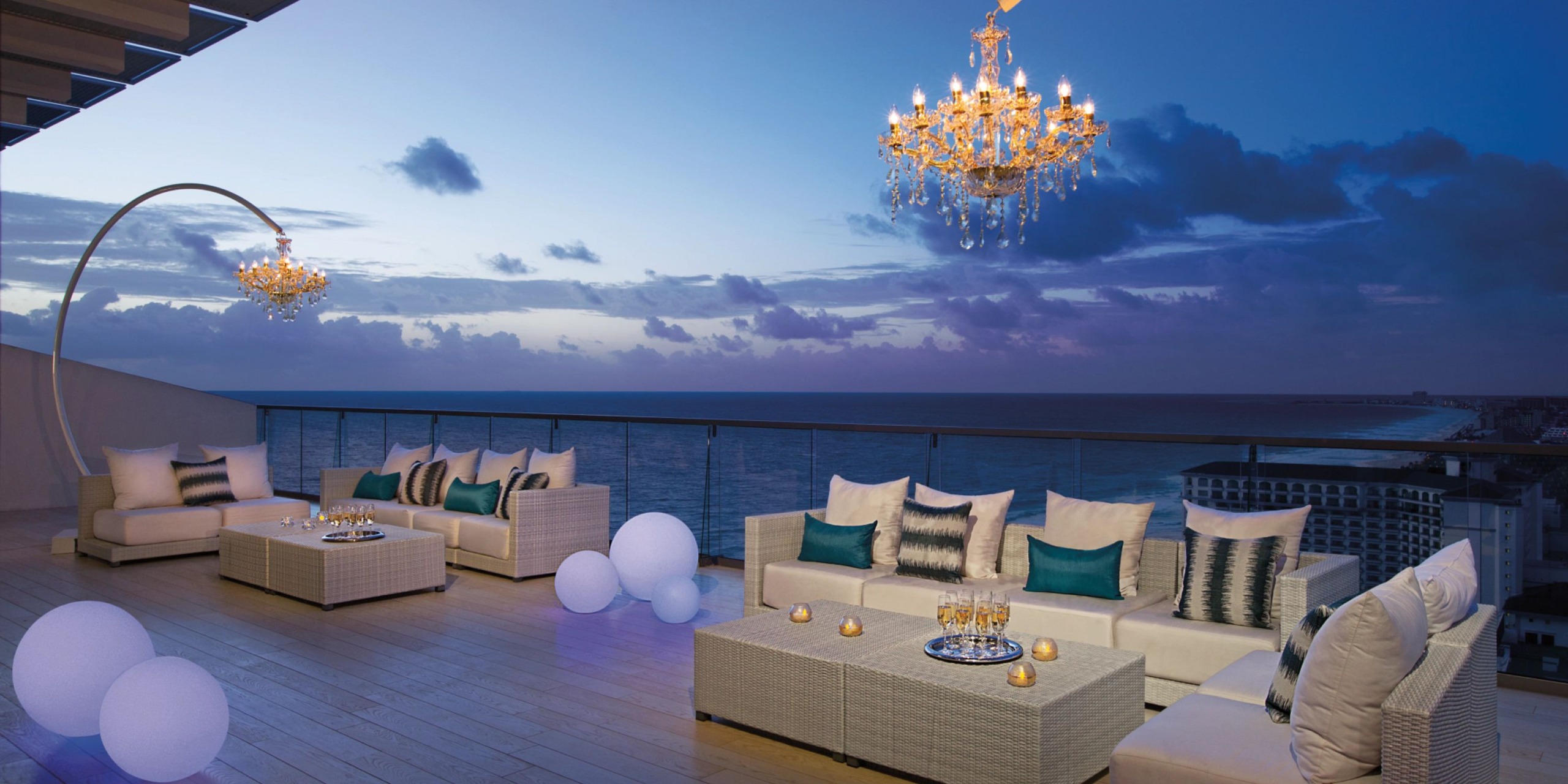 Experience the Secrets the Vine Cancun's rooftop lounge at sunset, complete with sleek modern white sofas adorned with teal cushions, two elegant chandeliers, a stylish glass railing, and breathtaking ocean views.