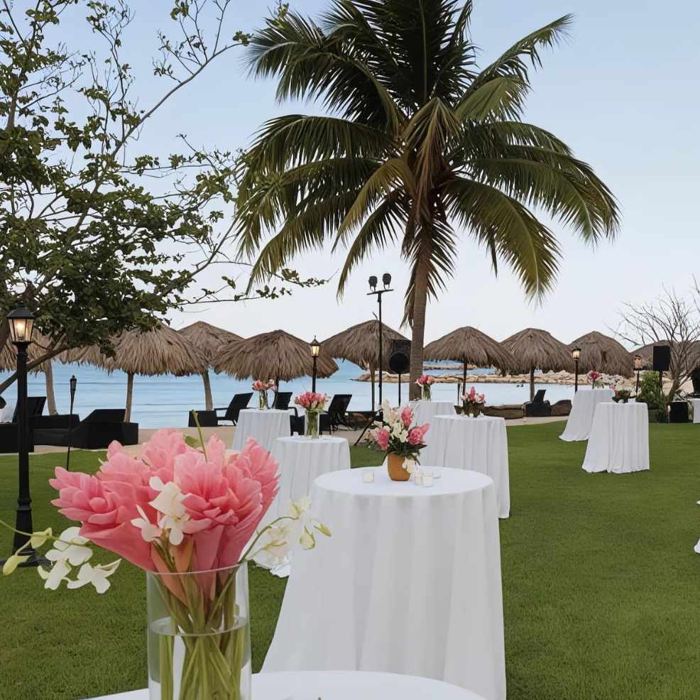 Outdoor event setup on a lawn with round tables draped in white tablecloths, pink floral centerpieces, and tiki umbrellas in the background near a beach with palm trees.