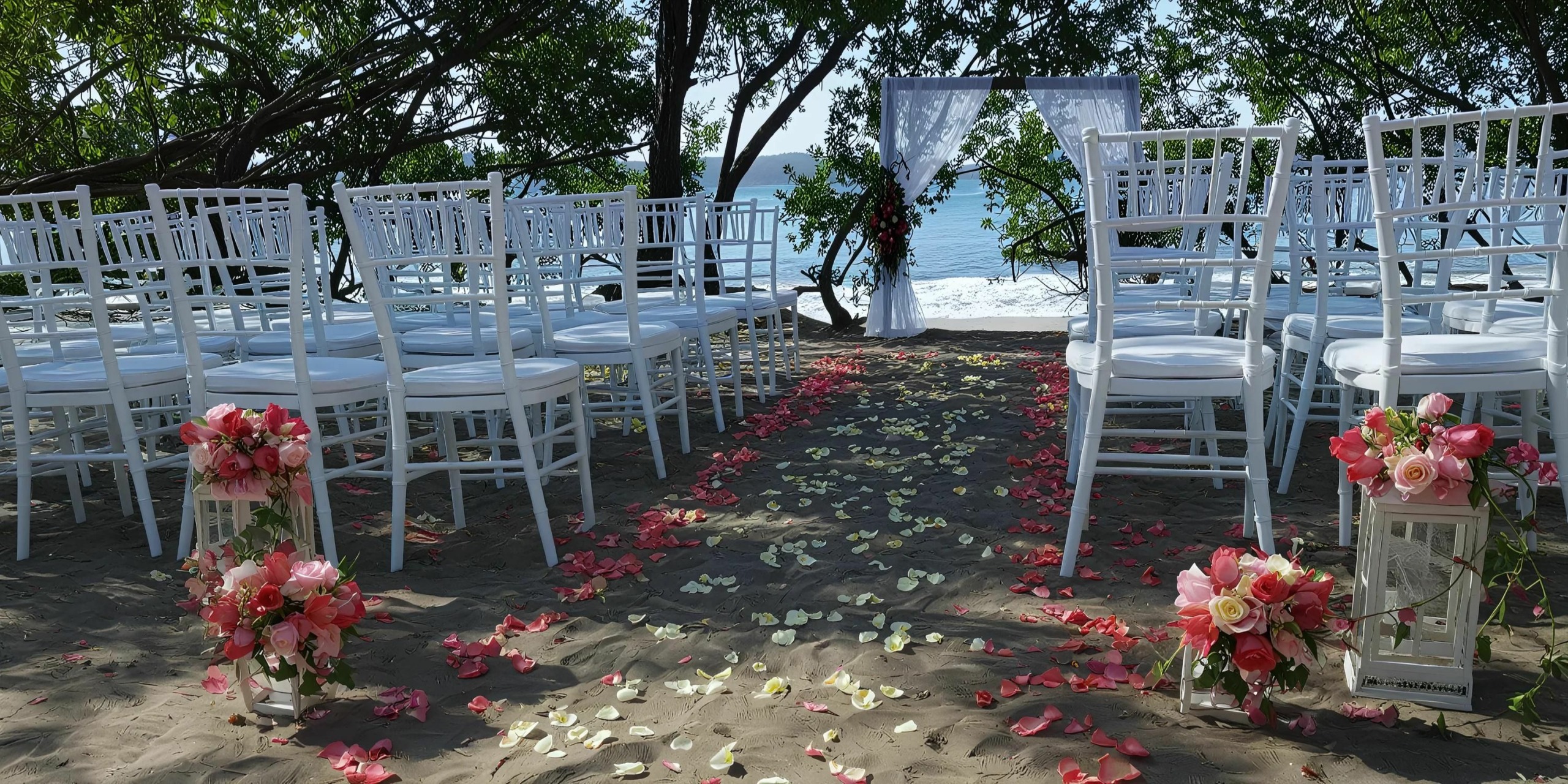 Outdoor beach wedding setup with white chairs arranged in rows, an aisle lined with red and white rose petals, and a white arch decorated with fabric in the background, overlooking the ocean.