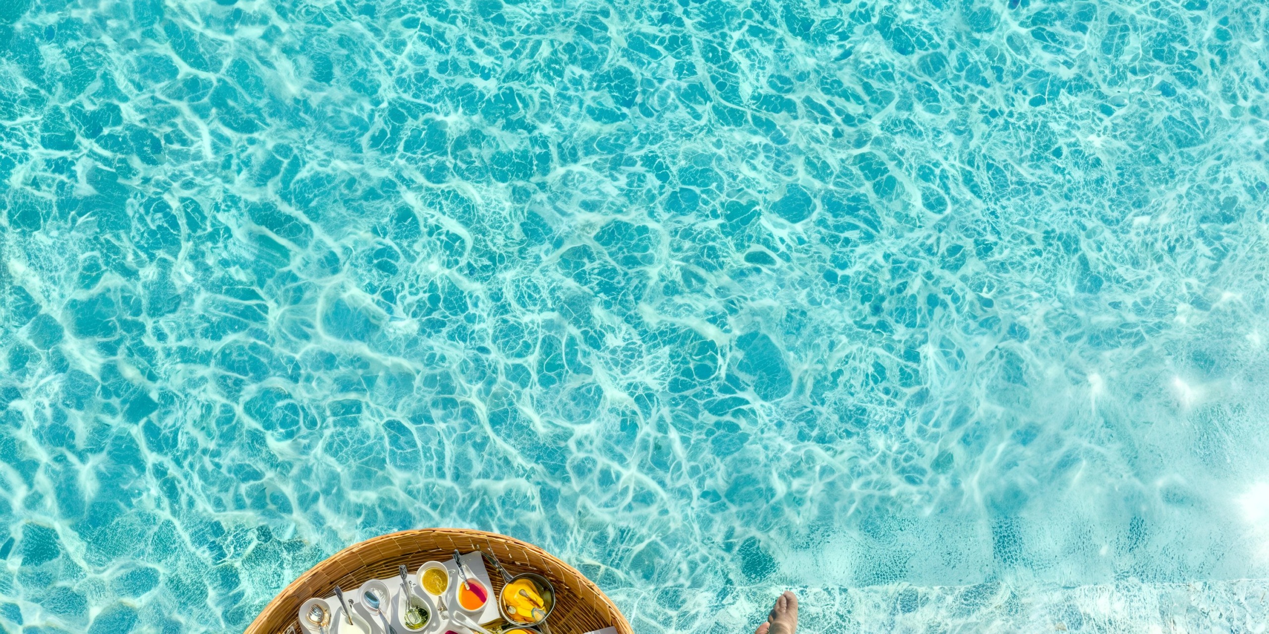 The Marival Armony floating breakfast tray, laden with a variety of foods and drinks, rests on a turquoise swimming pool.