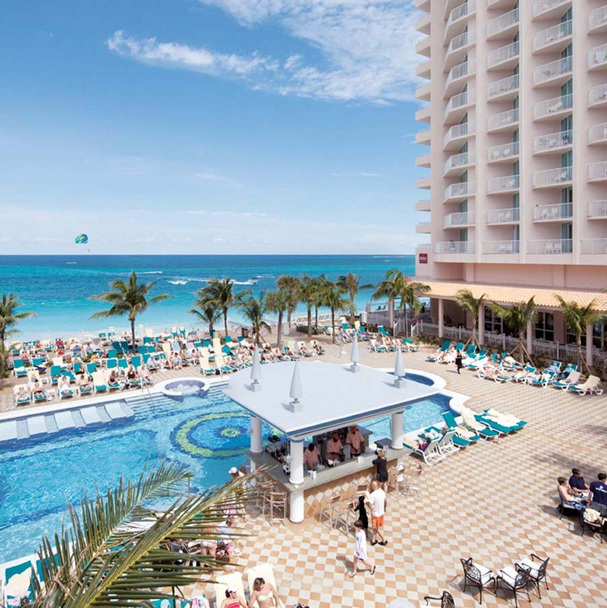 A destination wedding at Hotel Riu Palace Paradise Island with people sunbathing and a parasailer over the ocean, adjacent to a high-rise hotel.