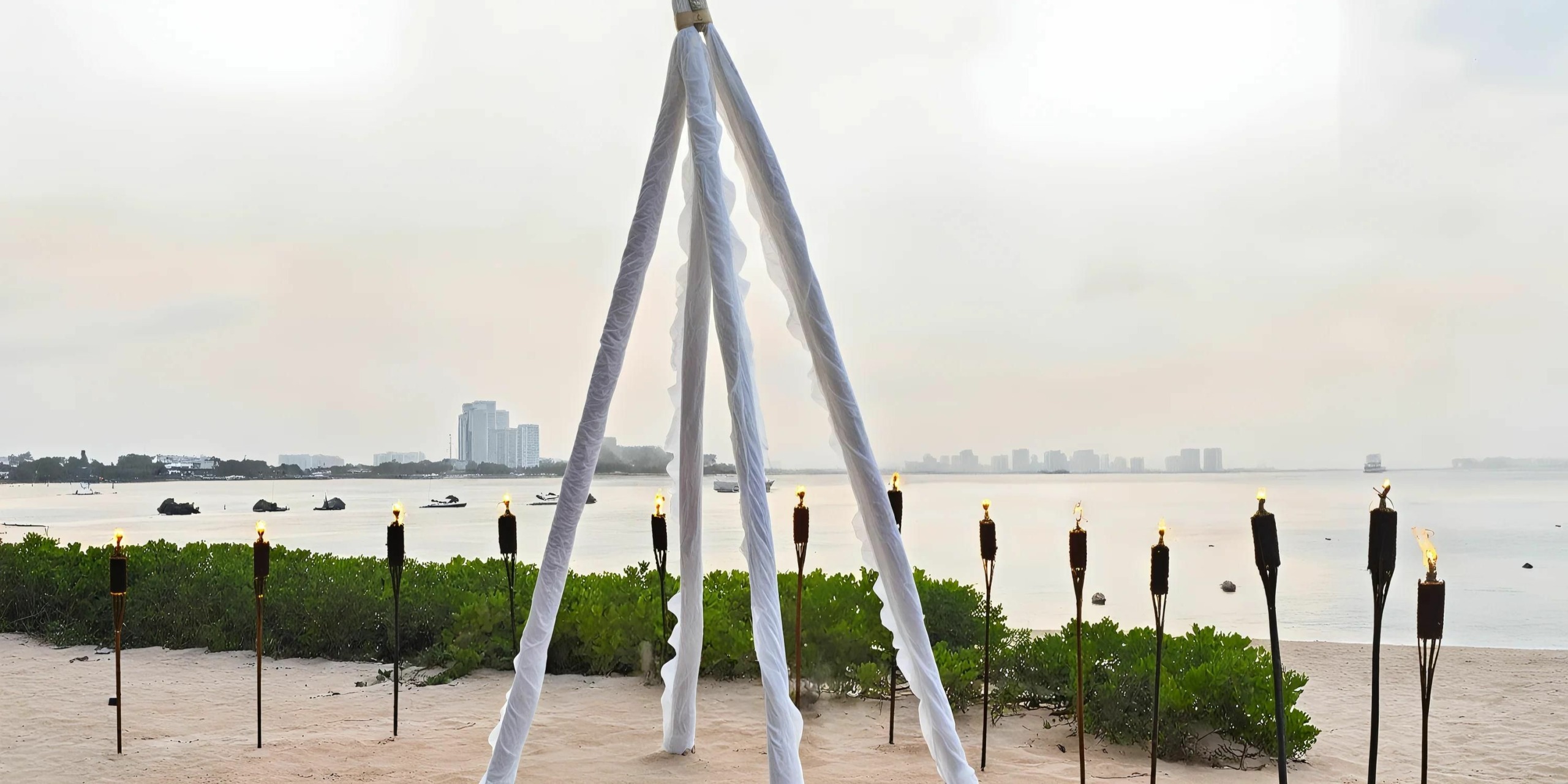 At Dreams Sands Cancun, an airy triangular structure draped with white fabric sits on a sandy beach, surrounded by tiki torches, with a cityscape visible in the distance across calm water.