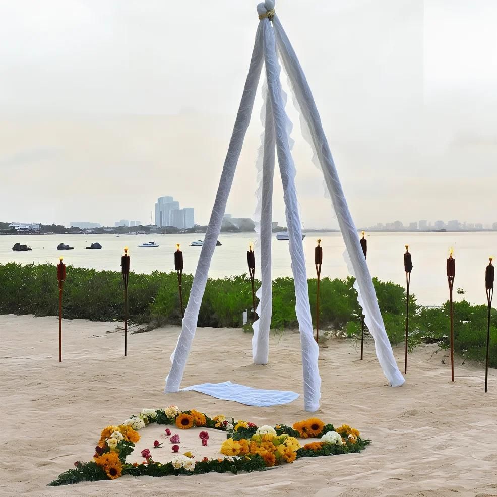 A beach setting with a white fabric canopy surrounded by torches. A blanket lies under the canopy, and a heart-shaped arrangement of flowers is on the sand. Water and buildings are visible in the background.