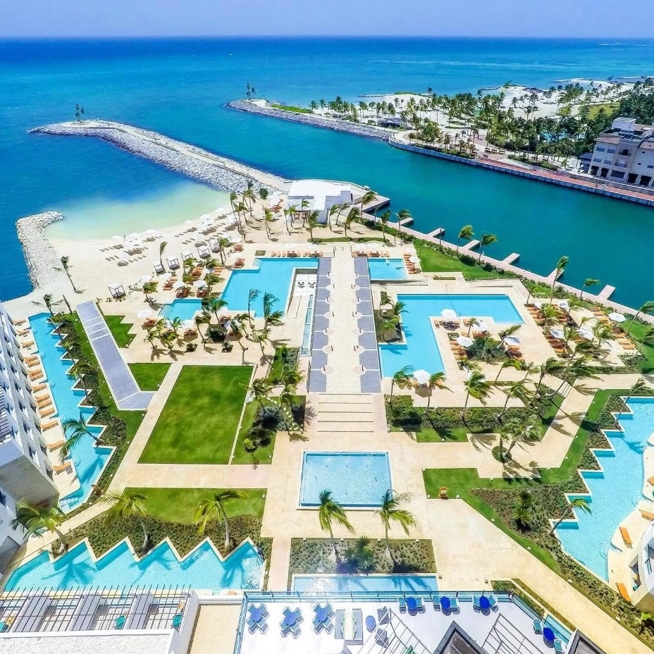 Aerial view of TRS Cap Cana Waterfront & Marina Hotel, popular for destination weddings, with multiple swimming pools and palm trees, adjacent to a beach and turquoise sea.