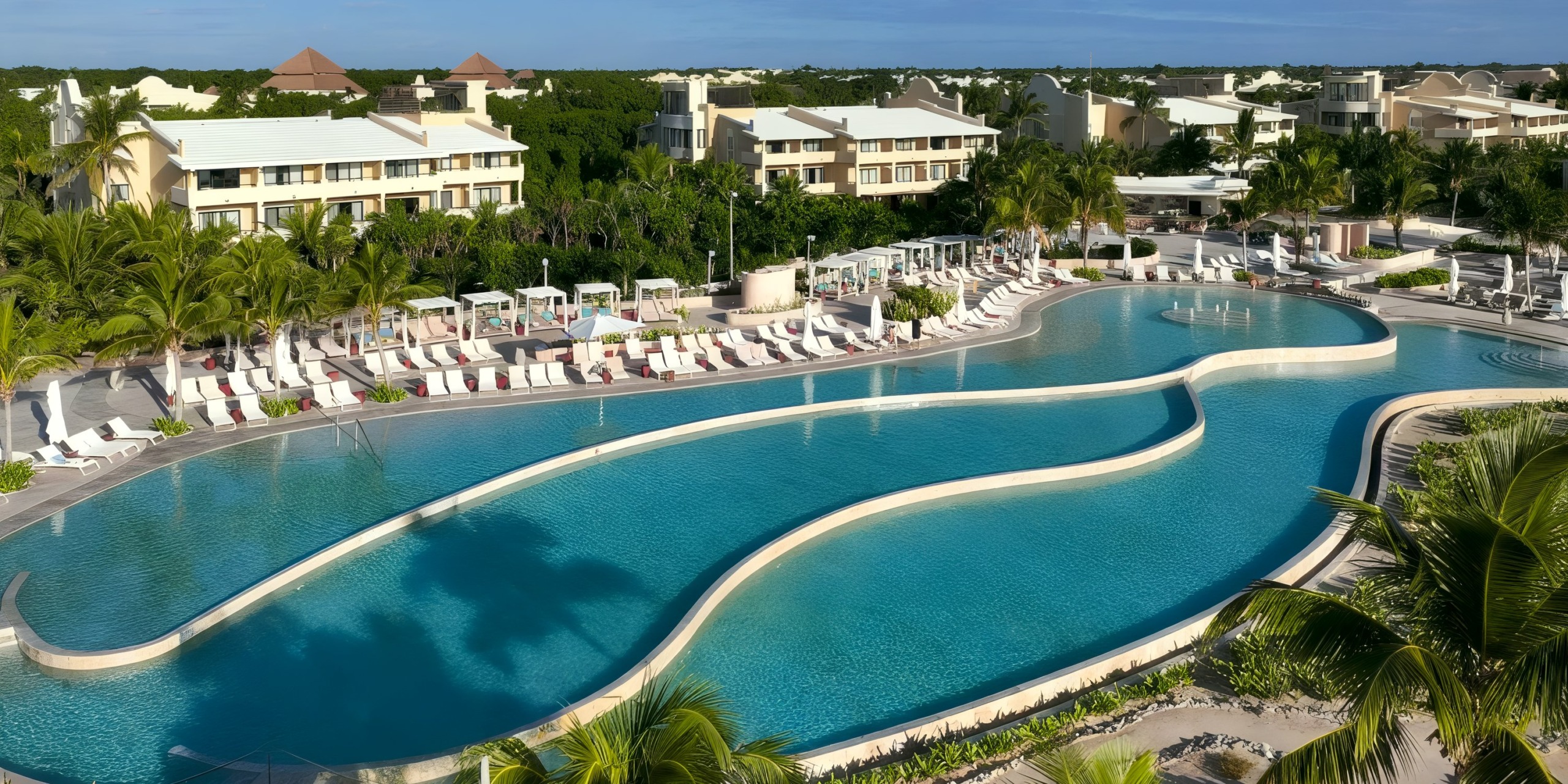 The TRS Yucatan features a sprawling curving outdoor pool flanked by comfortable lounge chairs and lush greenery, set against the backdrop of several buildings beneath a clear blue sky.