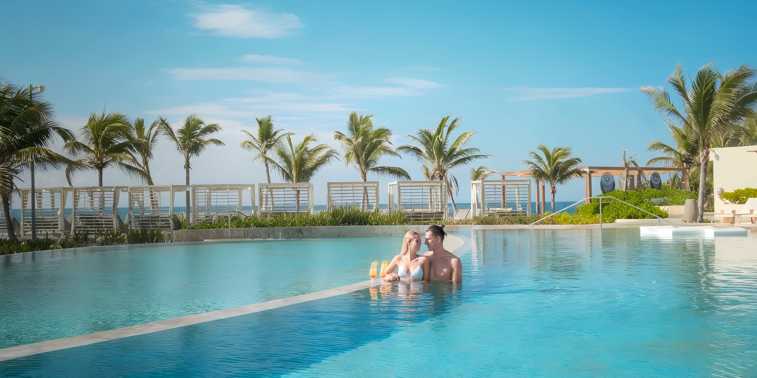Under the clear blue sky, a couple relaxes in the expansive outdoor TRS Yucatan pool, surrounded by palm trees and lounge chairs.