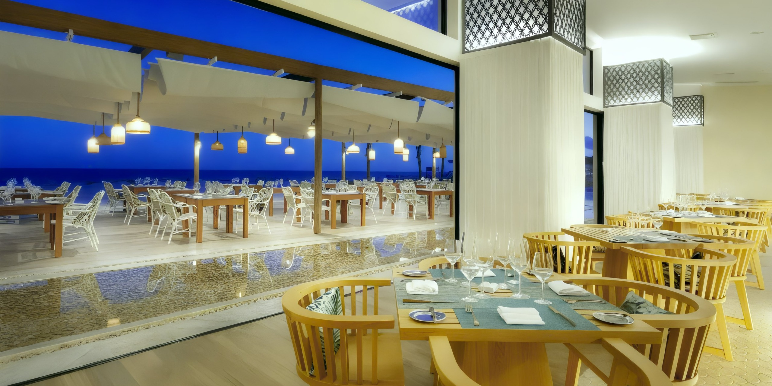 The TRS Yucatan features a contemporary dining area with wooden tables and chairs, which opens up to an outdoor patio. This patio, adorned with umbrellas, offers a stunning view of a body of water beneath a deep blue sky.