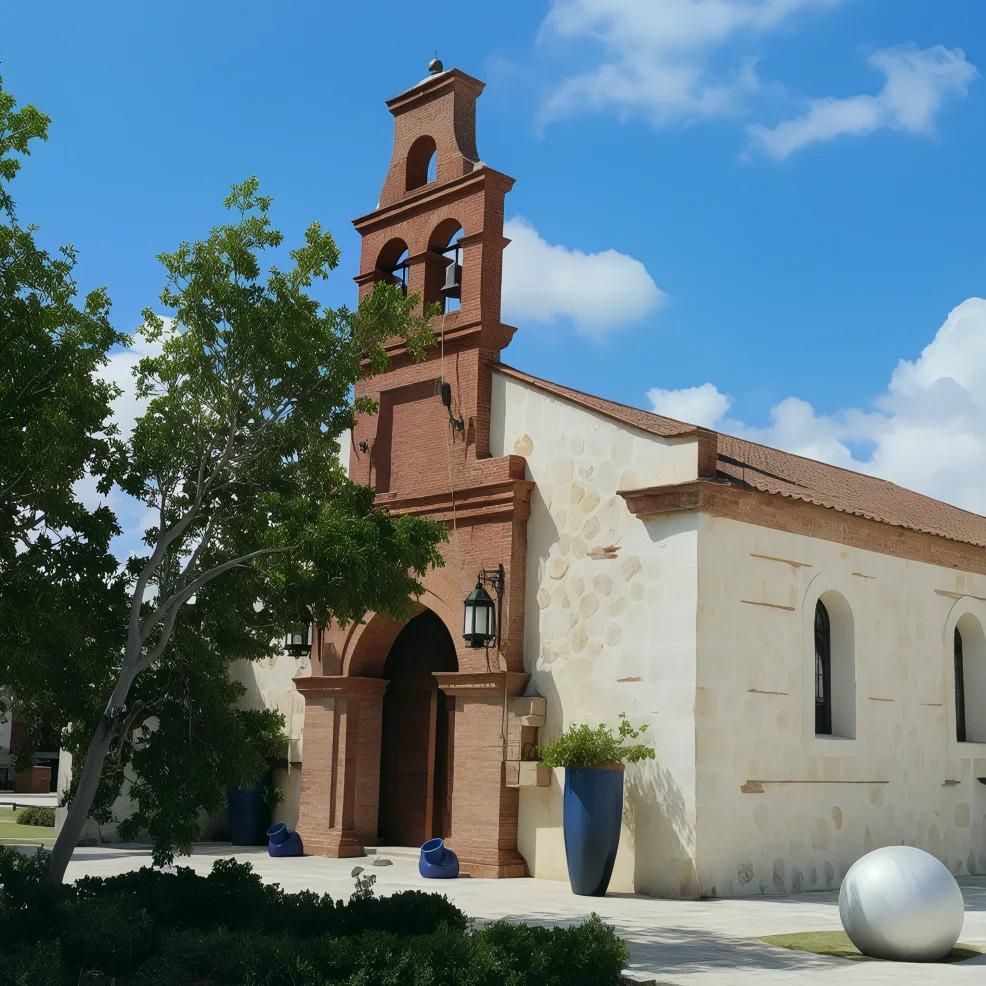 A small, beige chapel with a brick bell tower, surrounded by trees and large decorative pots under a blue sky.