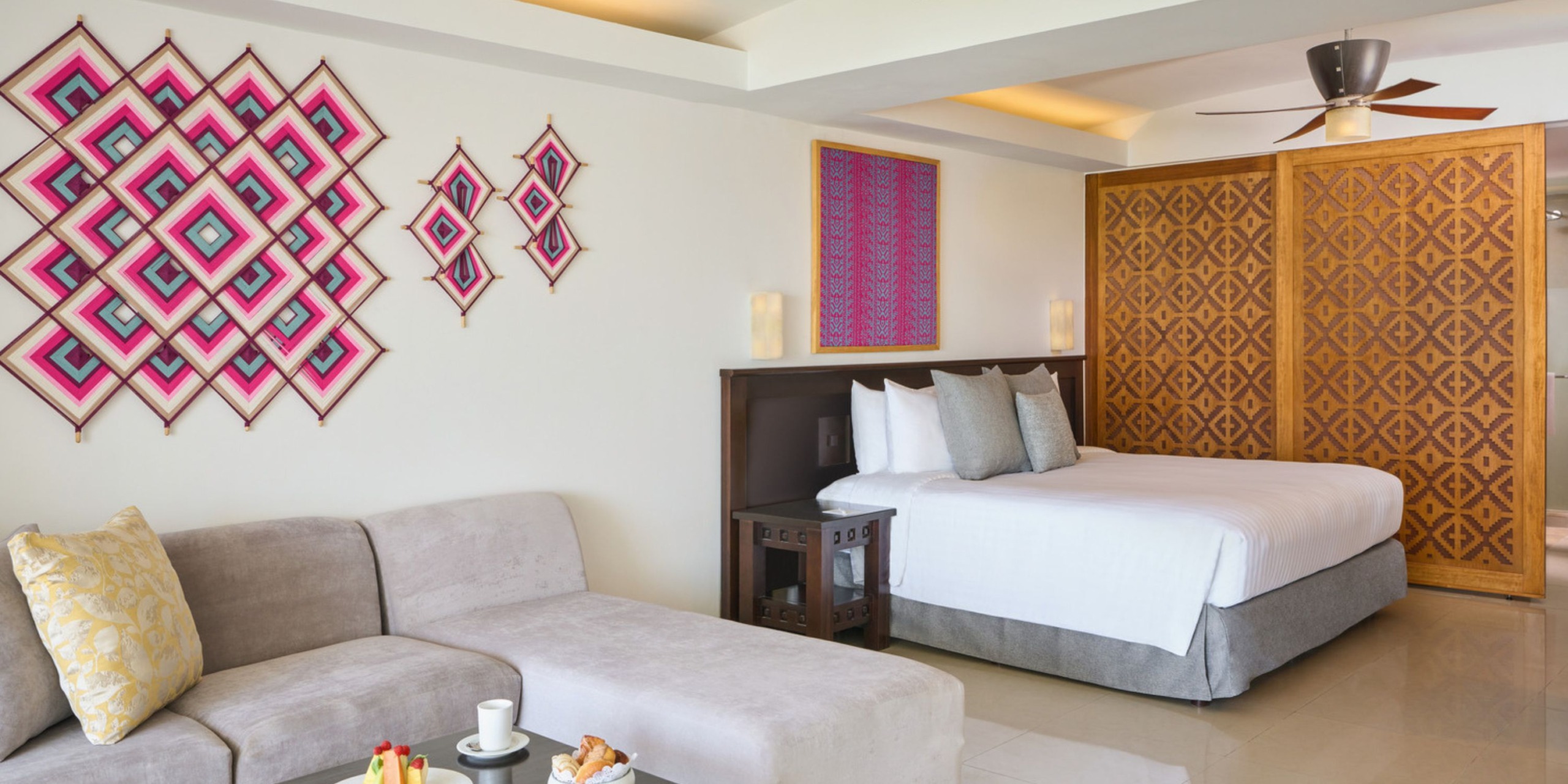 Experience a stay at the Wyndham Alltra Vallarta in a hotel room featuring a double bed, gray sofa, decorative wall patterns, and a ceiling fan. Enjoy a small table set with a refreshing drink and an assortment of snacks.