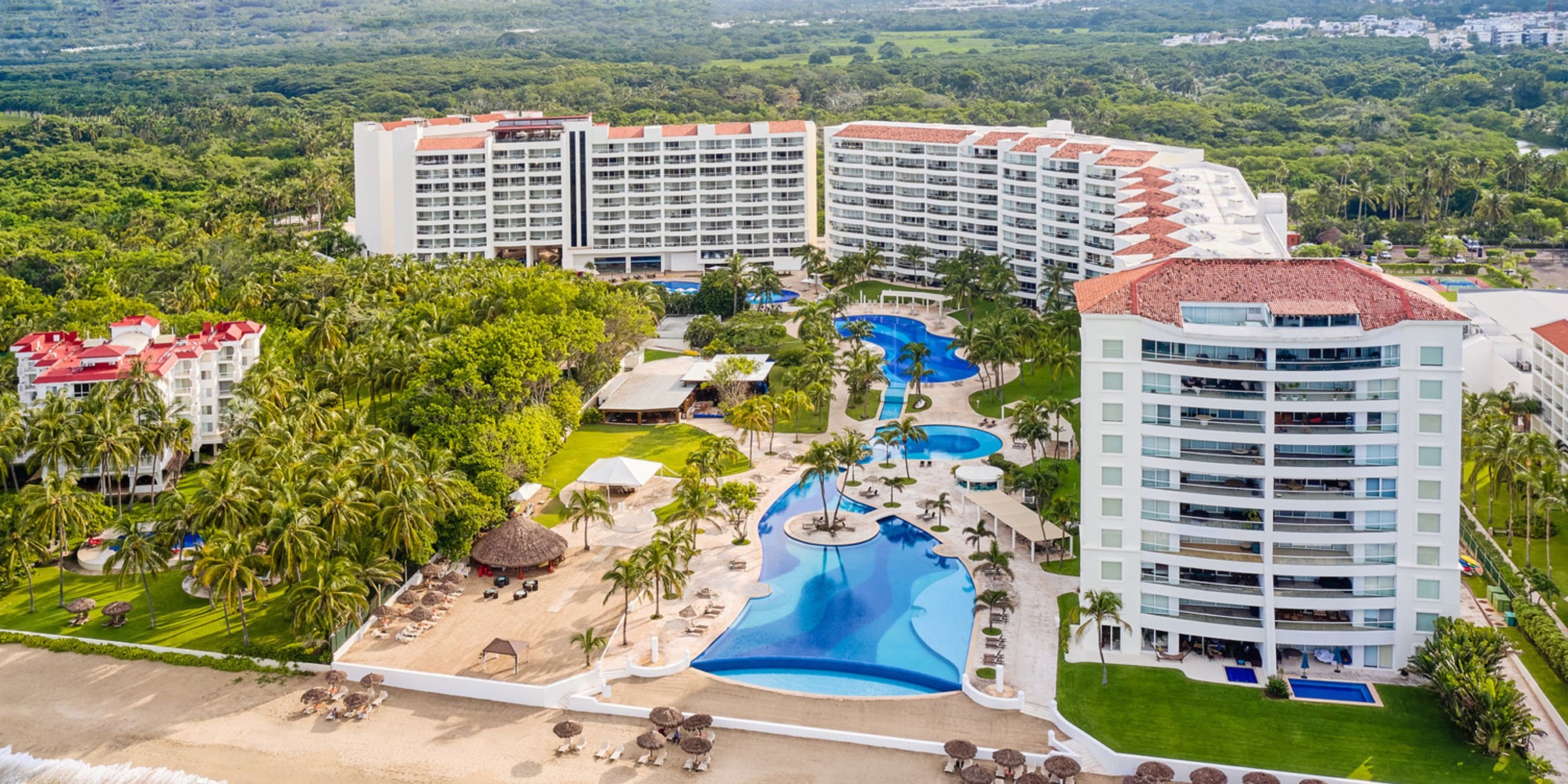 Aerial view of the Wyndham Alltra Vallarta, a beachfront hotel featuring multiple white buildings, shimmering blue swimming pools, and palm trees, all nestled amid lush greenery.
