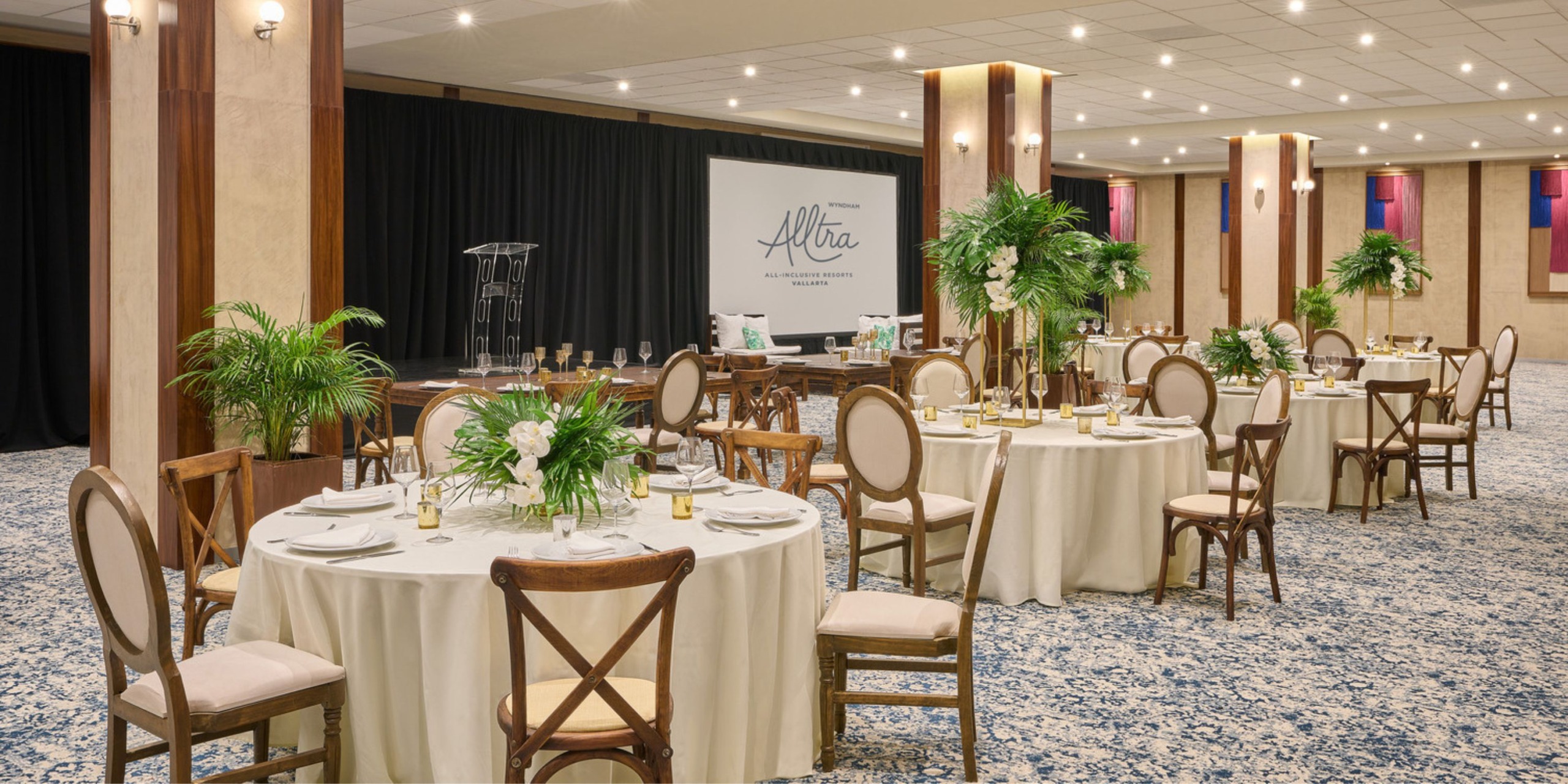 The Wyndham Alltra Vallarta offers an elegant event space featuring round tables with floral centerpieces. A stage equipped with a podium and scenic backdrop is readily visible. The venue's walls are decorated with classic columns and lush plants, complemented by a patterned carpeted floor.