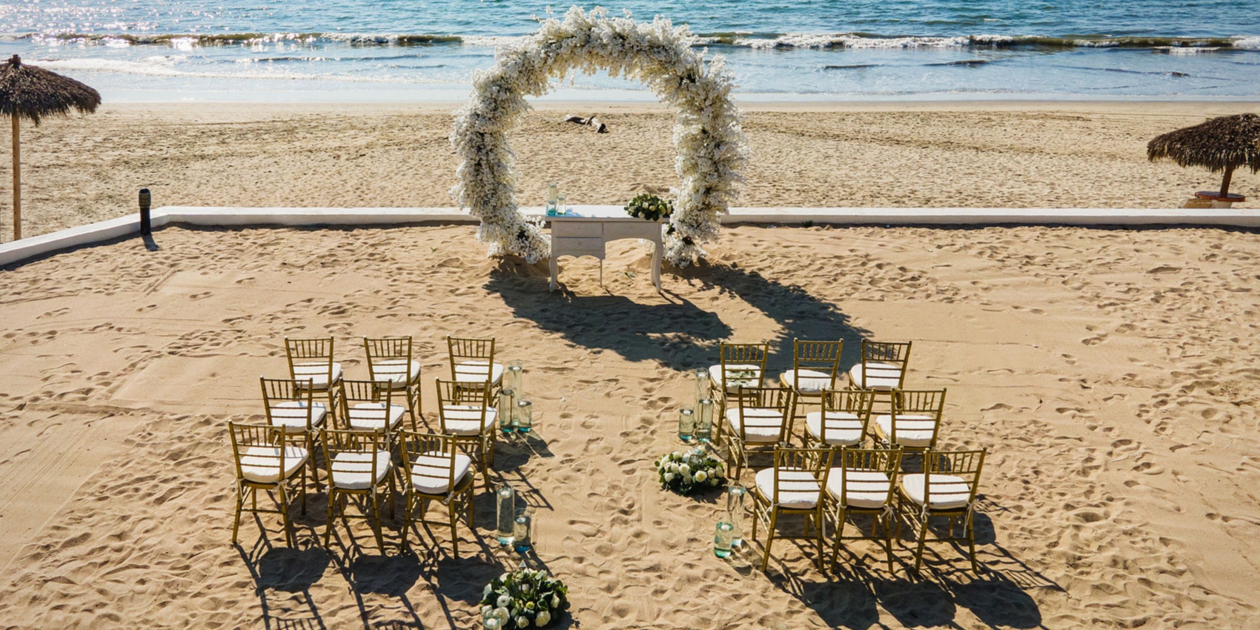 Experience an elegant beach wedding at Wyndham Alltra Vallarta, complete with a floral arch, gold chairs, and floral arrangements overlooking the ocean on a sandy shore.