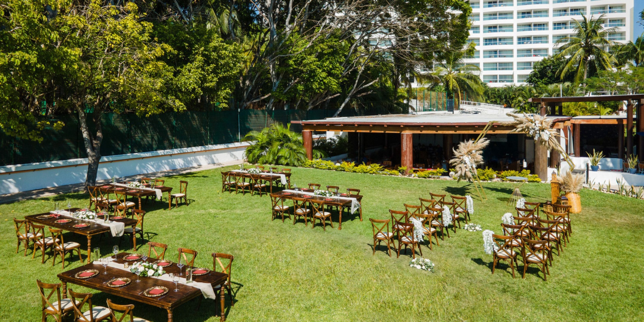The setup for the outdoor event features Wyndham Alltra Vallarta's wooden tables and chairs arranged on a grassy area, with trees surrounding and a building visible in the background.