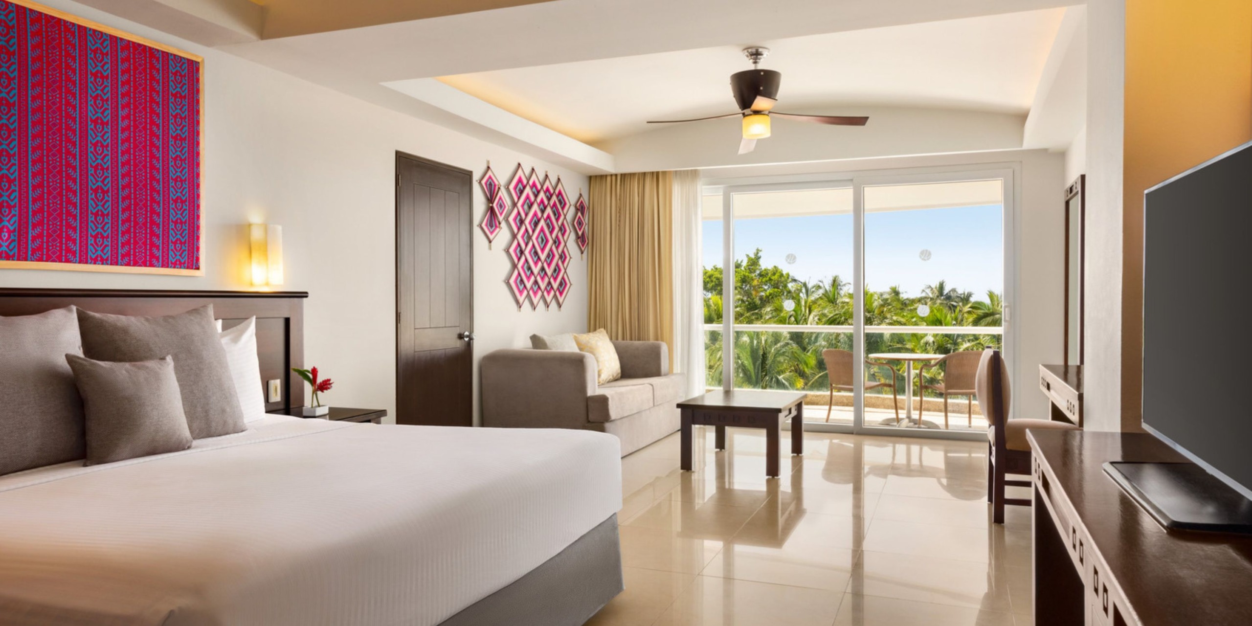Room at Wyndham Alltra Vallarta featuring a large bed, cozy sofa, coffee table, and television. A ceiling fan overhead complements the glass doors opening onto a balcony with views of palm trees. The walls are adorned with vibrant artwork.