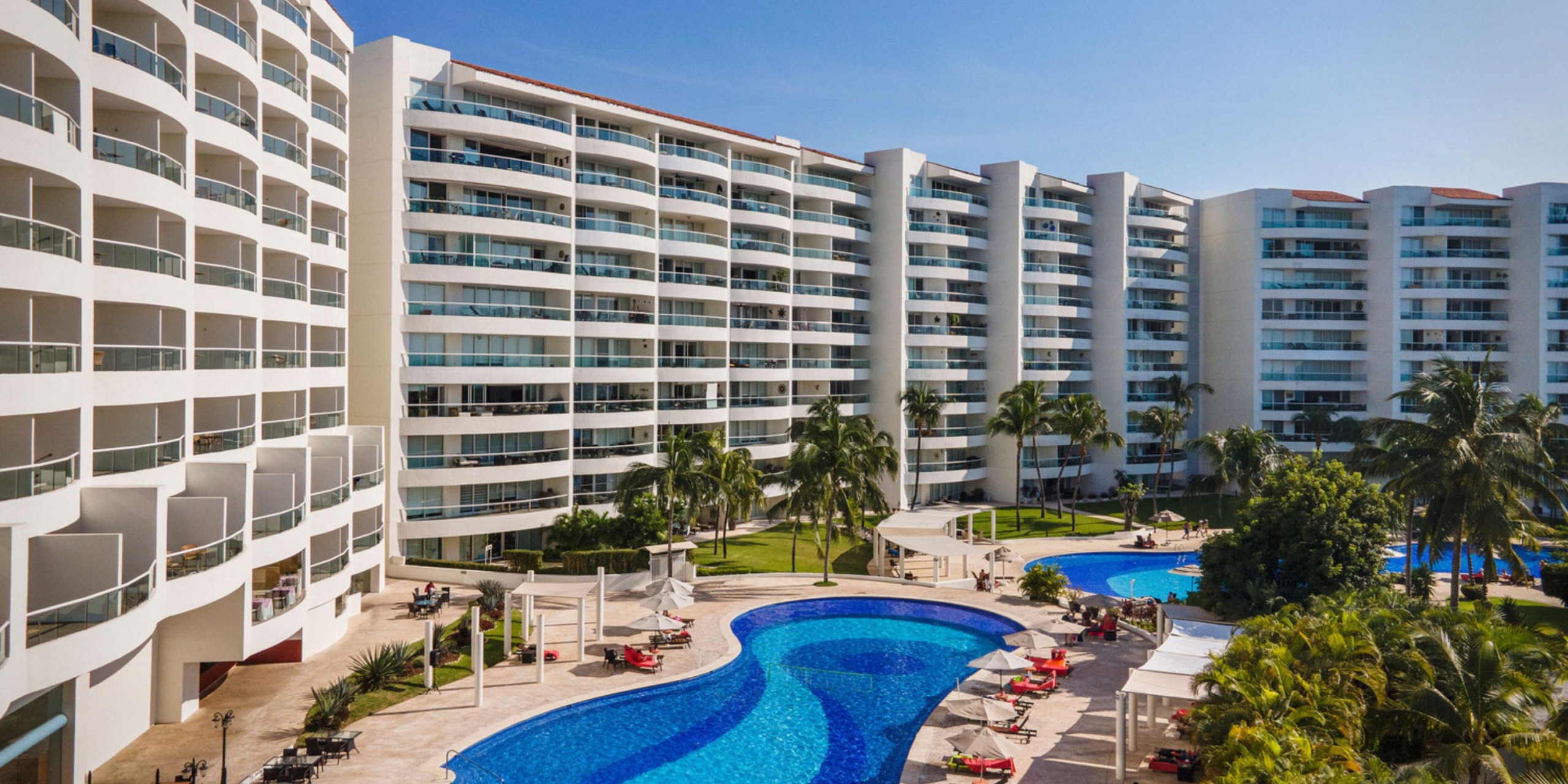 Aerial view of Wyndham Alltra Vallarta, a contemporary hotel with multiple floors, showcasing palm trees, a spacious swimming pool, and sun loungers neatly arranged around the pool area under clear blue skies.