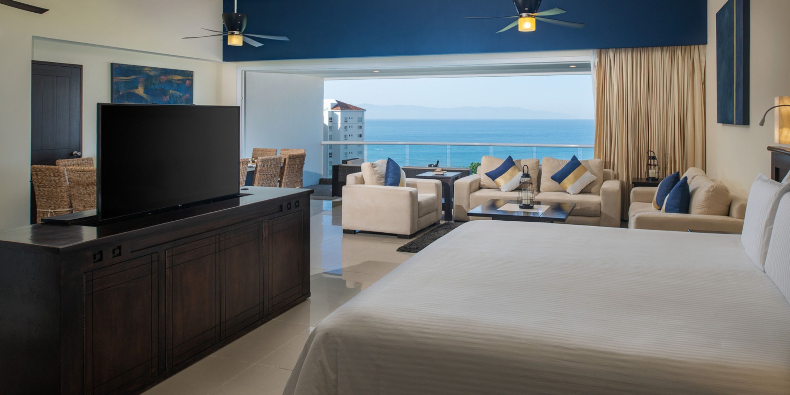 Enjoy a spacious room at the Wyndham Alltra Vallarta, featuring a comfortable bed, flatscreen TV, cozy seating area, and a balcony with stunning views of the ocean and distant buildings.