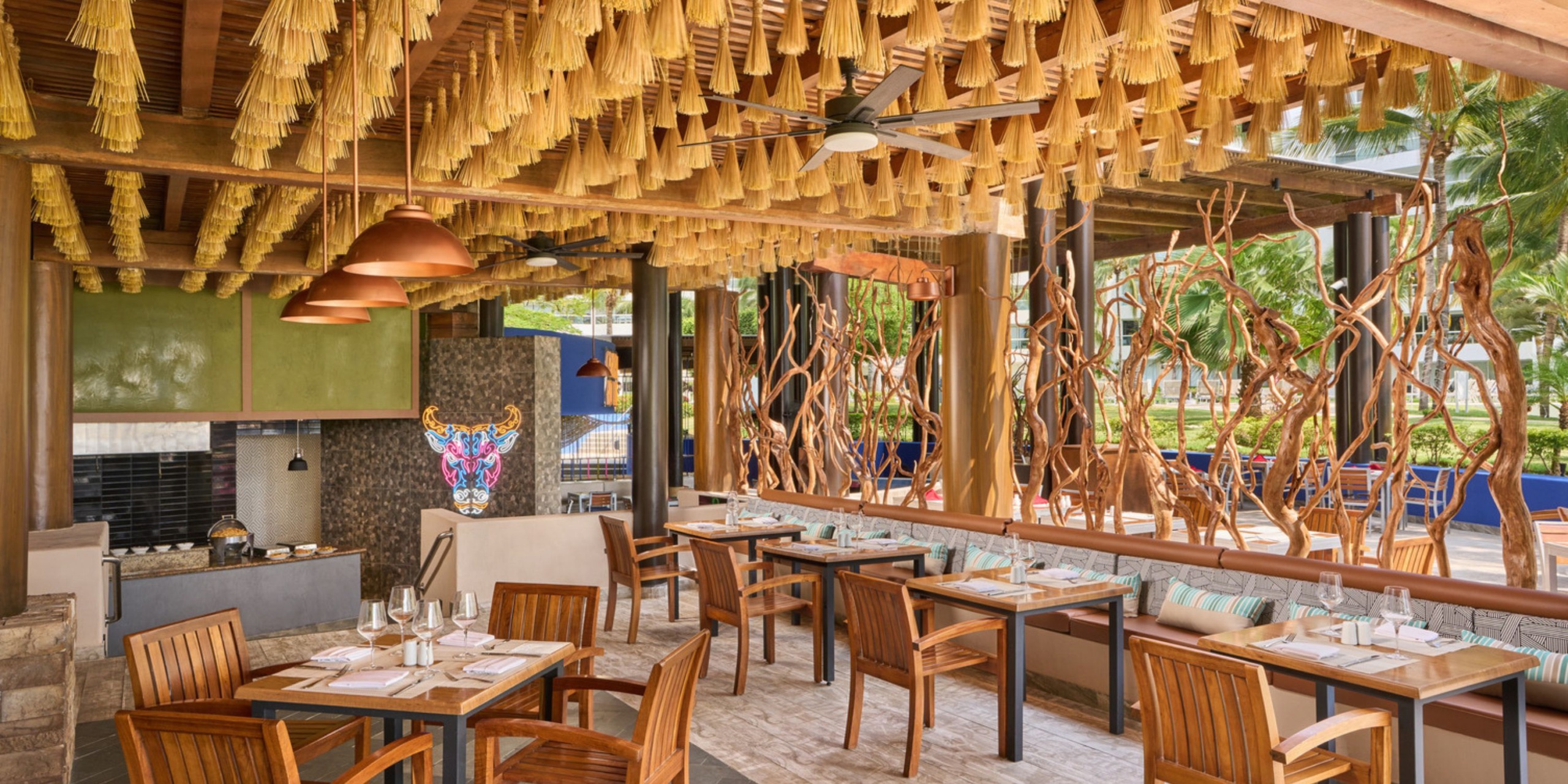 The outdoor dining area at Wyndham Alltra Vallarta features wooden tables and chairs beneath a ceiling adorned with decorative hangings, surrounded by lush greenery and artistic elements in the background.