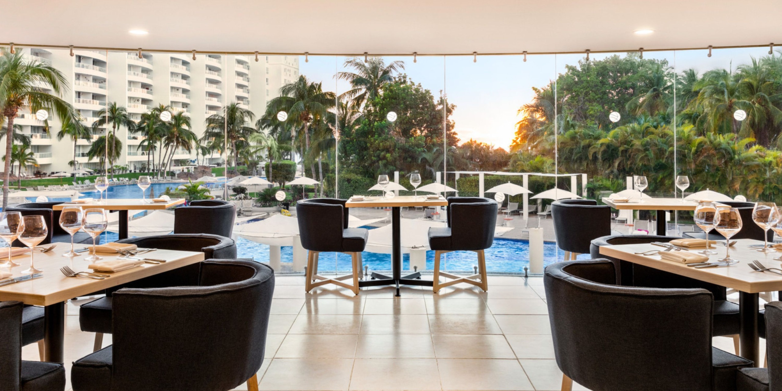 The dining area at Wyndham Alltra Vallarta features set tables and chairs that overlook the pool and palm trees, with a view of a high-rise building and a stunning sunset in the background.