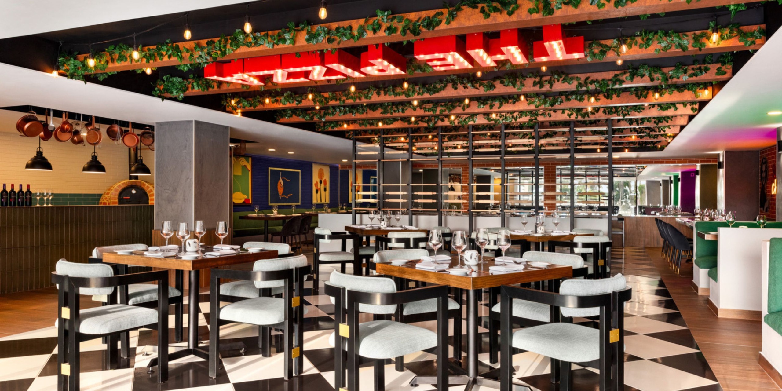 Wyndham Alltra Vallarta offers a chic dining experience with a black-and-white checkered floor, wooden tables ready for guests, chairs featuring green cushions, and a ceiling beautifully decorated with greenery and lights.