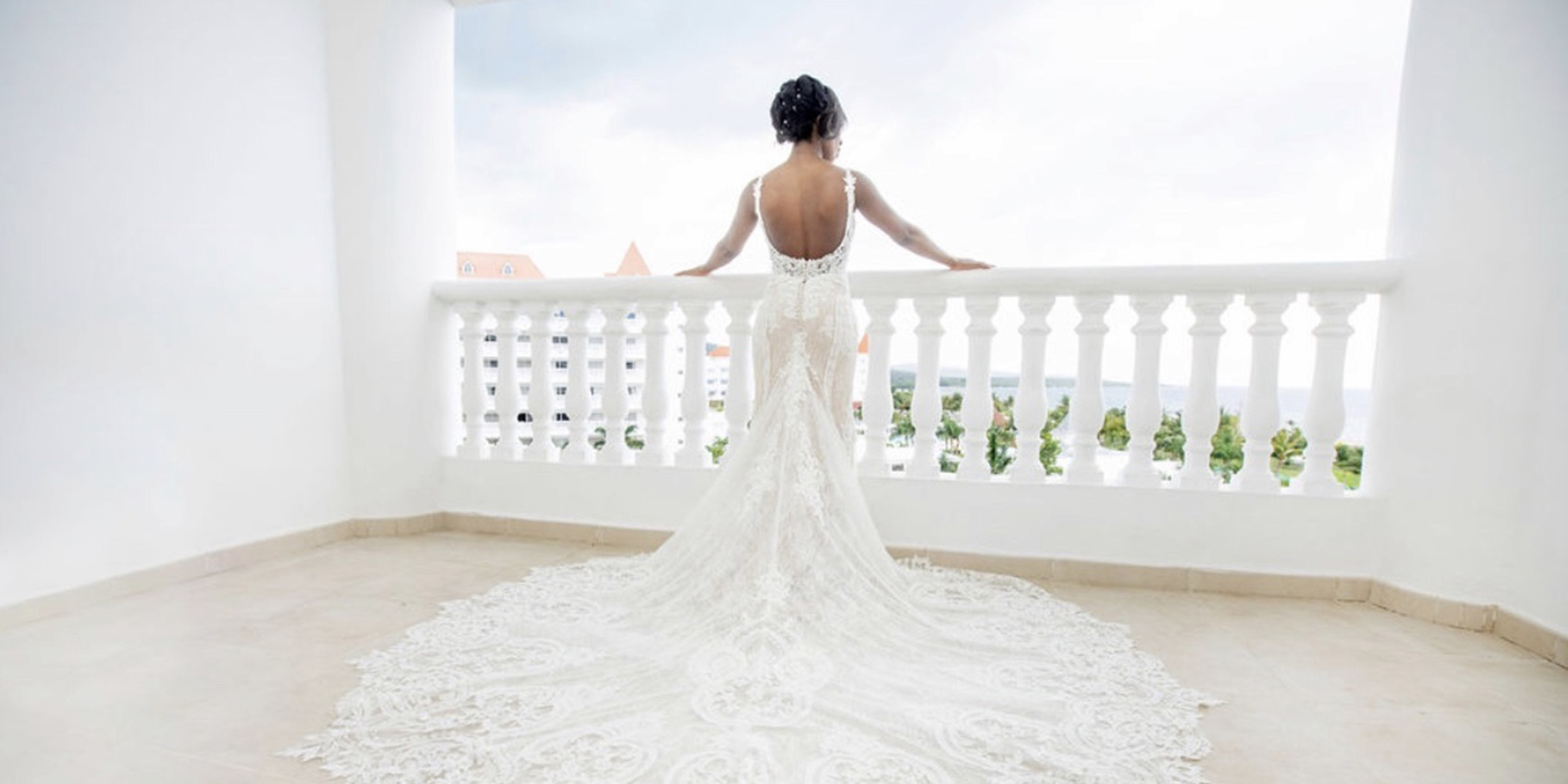 A bride in the elegant Wokie and James wedding dress, adorned with intricate lace, stands on a balcony, facing away, overlooking a scenic view.