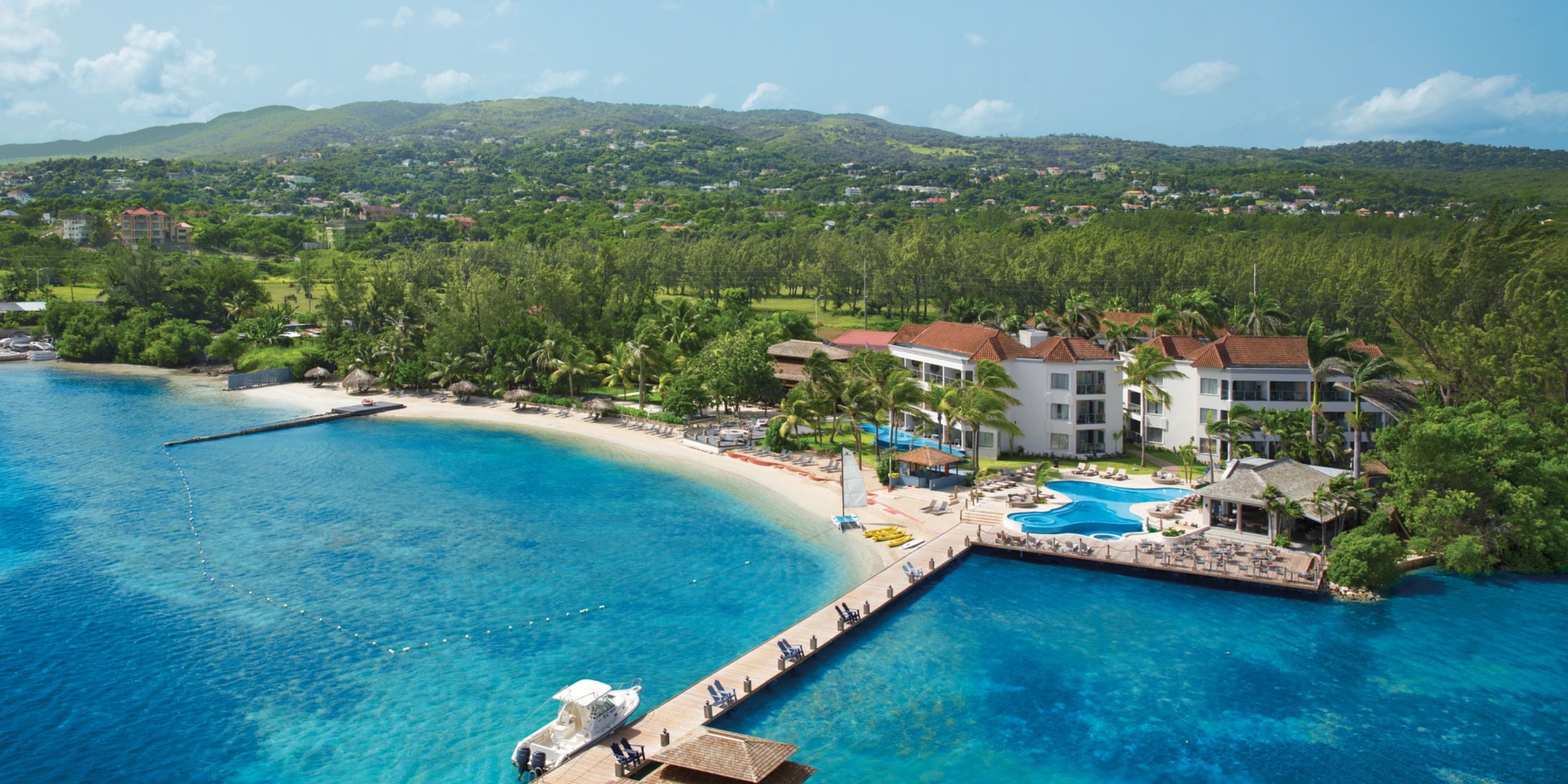 Aerial view of Zoetry Montego Bay, a resort by the sea featuring a long pier, clear blue water, a sandy beach lined with palm trees, and surrounded by lush green hills.