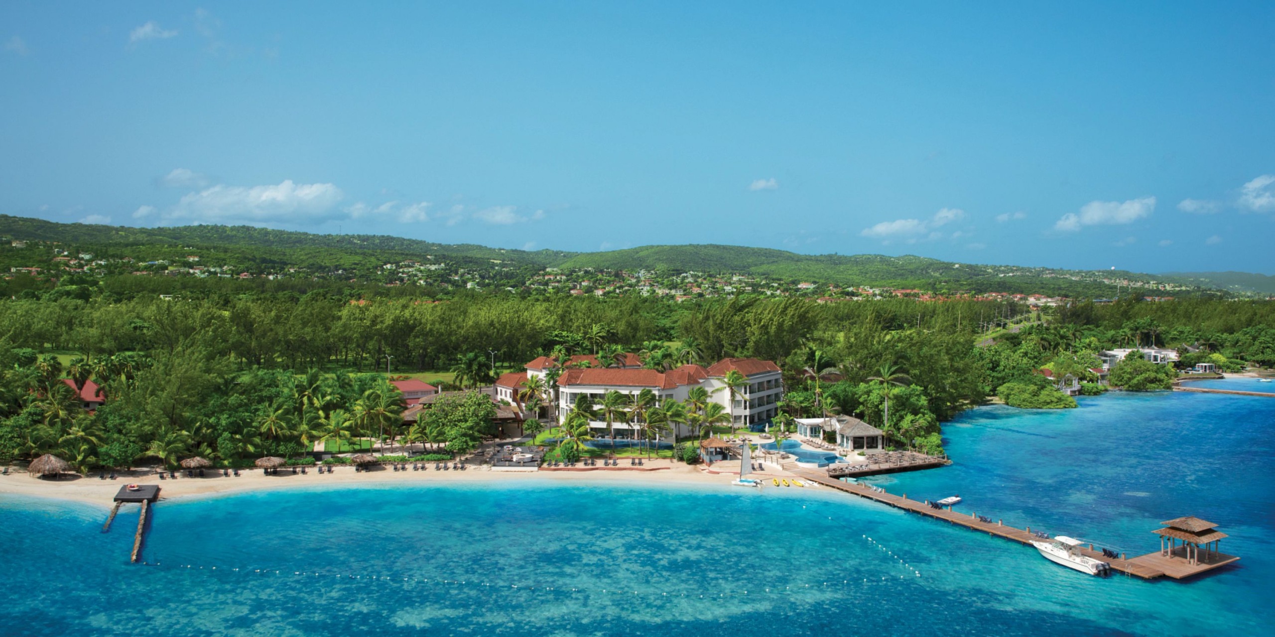 Aerial view of Zoetry Montego Bay, a coastal resort featuring a beach with clear turquoise water, a pier, and lush greenery set against a backdrop of a clear blue sky.