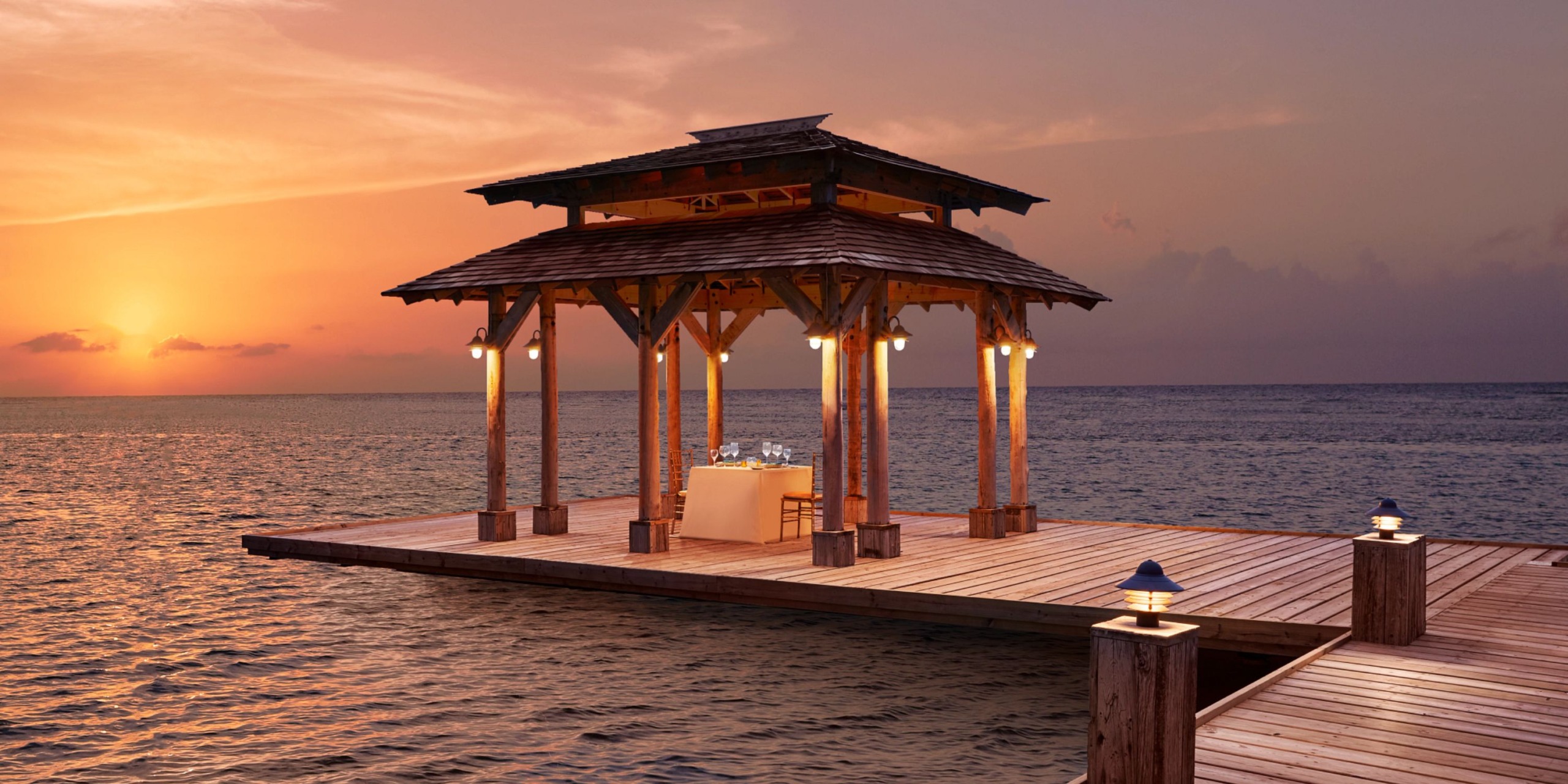At Zoetry Montego Bay, enjoy dining at a wooden gazebo located on a pier over the ocean, perfectly set against the backdrop of a sunset.