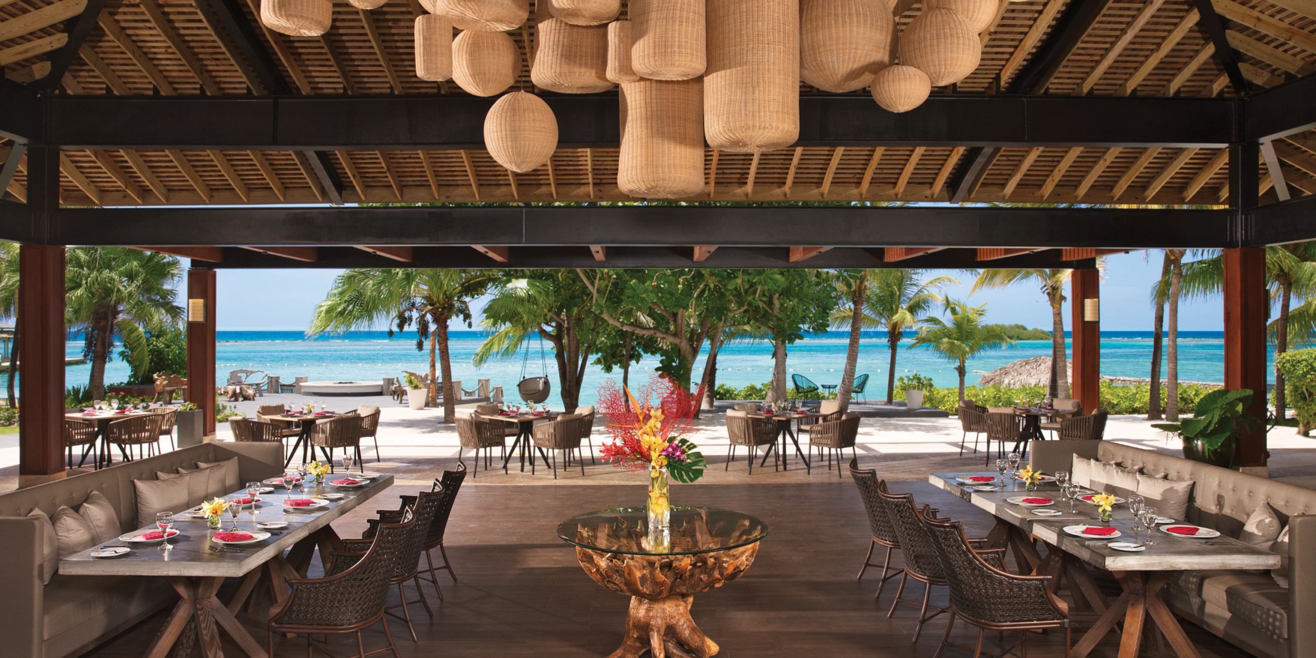 Experience beachfront dining at Zoetry Montego Bay, where wooden furniture, hanging lanterns, and stunning ocean views create the perfect ambiance. Each table is elegantly arranged with plates and red napkins.