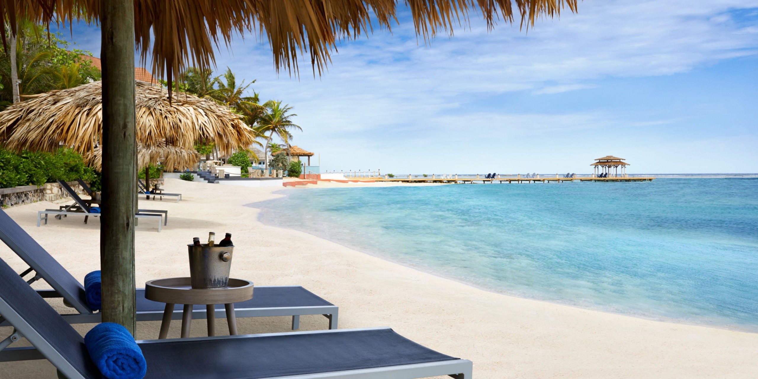 Experience the stunning Zoetry Montego Bay, where you can relax on a sandy beach complete with lounge chairs and palm-thatched umbrellas, enjoy refreshments from a small table with a bucket of drinks, and gaze at the ocean view and wooden dock beneath a clear blue sky.