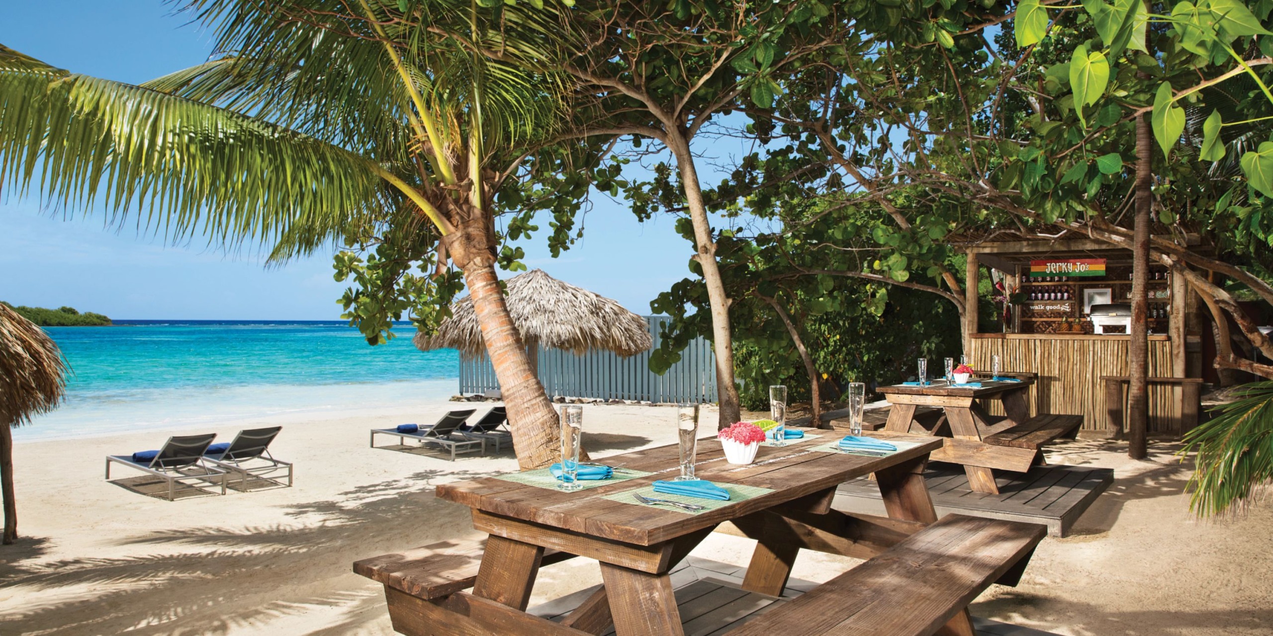 Zoetry Montego Bay offers wooden picnic tables nestled under palm trees on a sandy beach, complete with an ocean view and a quaint beach bar to the right.