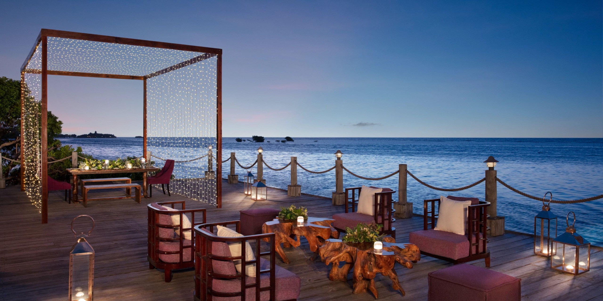 Zoetry Montego Bay offers an outdoor dining area by the sea at dusk, showcasing wooden tables and chairs beneath a canopy adorned with string lights, all set against a stunning view of the ocean.