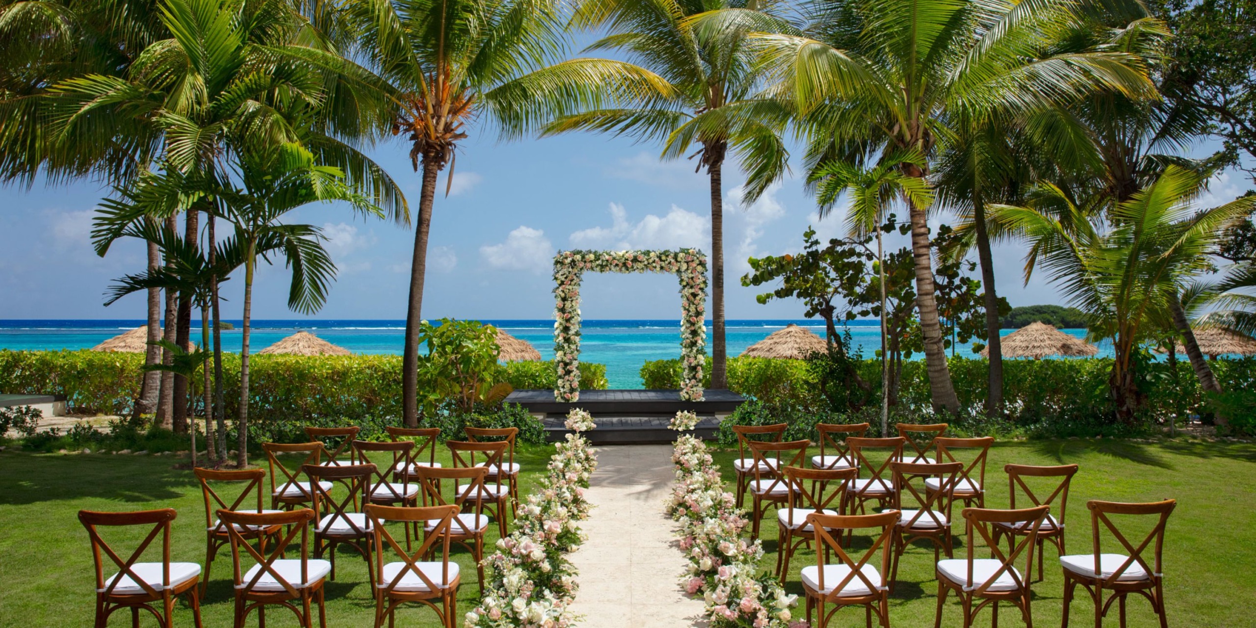 Experience a breathtaking outdoor wedding setup at Zoetry Montego Bay, where wooden chairs are elegantly arranged facing a beautifully adorned floral arch. Surrounded by swaying palm trees, your ceremony boasts a stunning ocean backdrop under the expansive blue sky.