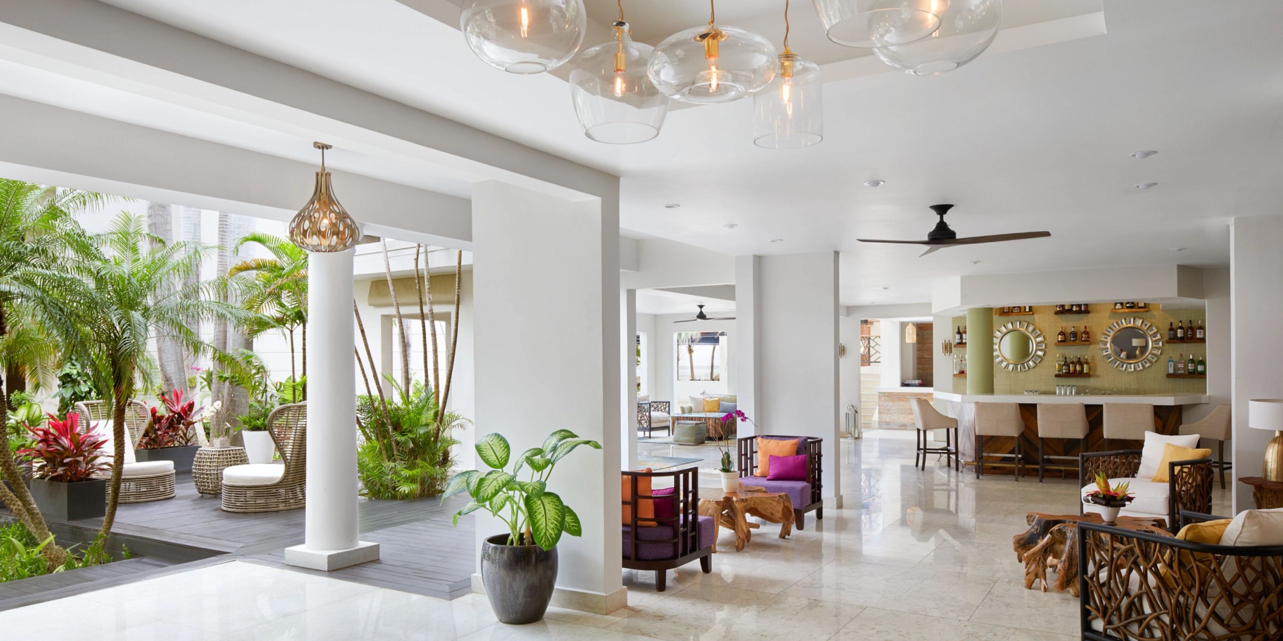 The Zoetry Montego Bay features a spacious modern lounge with white walls, tropical plants, wicker chairs, a bar area complete with stools, and a ceiling fan, all beautifully illuminated by pendant lights and natural light.