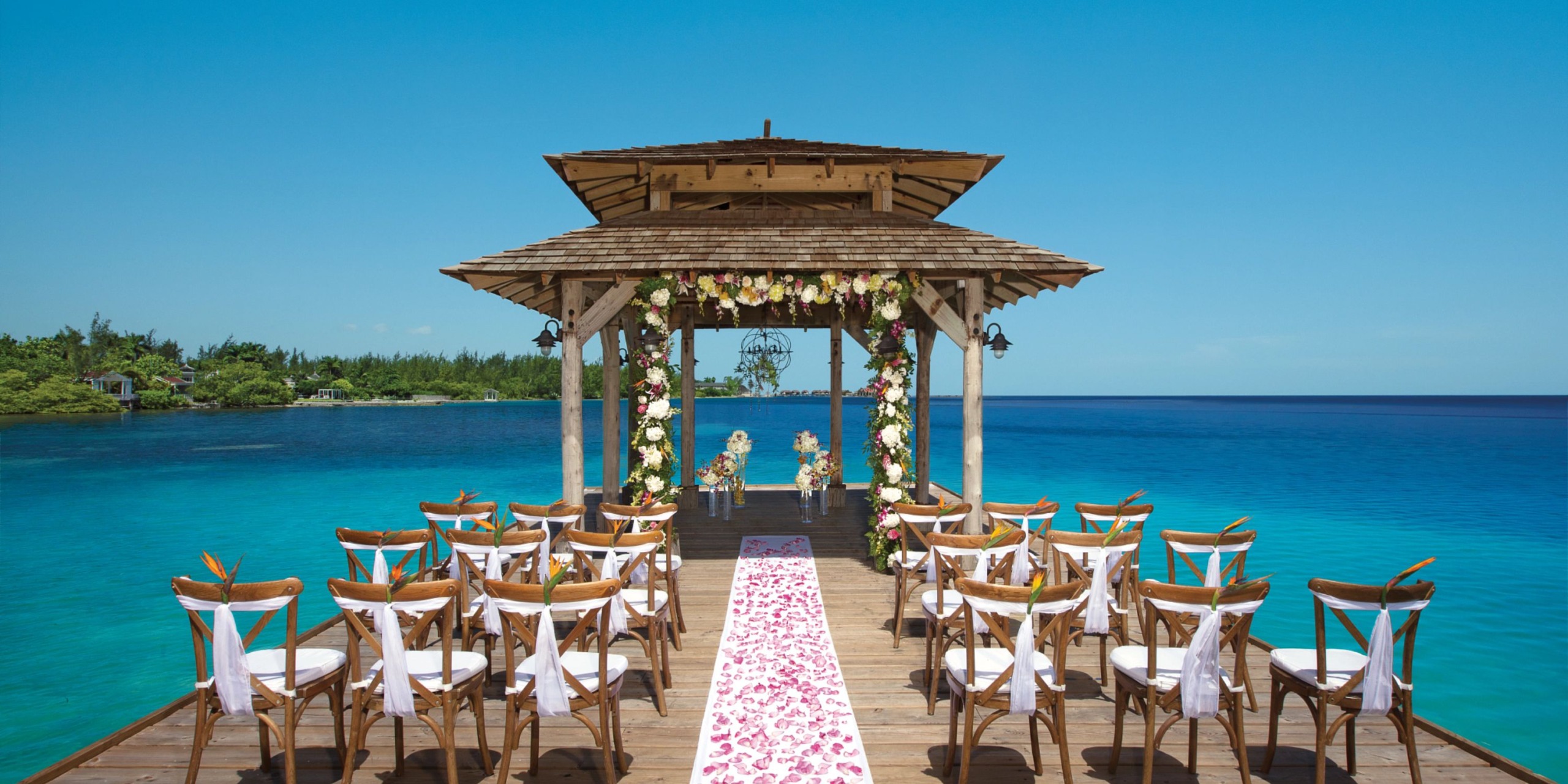 The ceremony setup at Zoetry Montego Bay includes a charming wooden pier with a flower-adorned gazebo, surrounded by rows of chairs facing the ocean under a clear blue sky.
