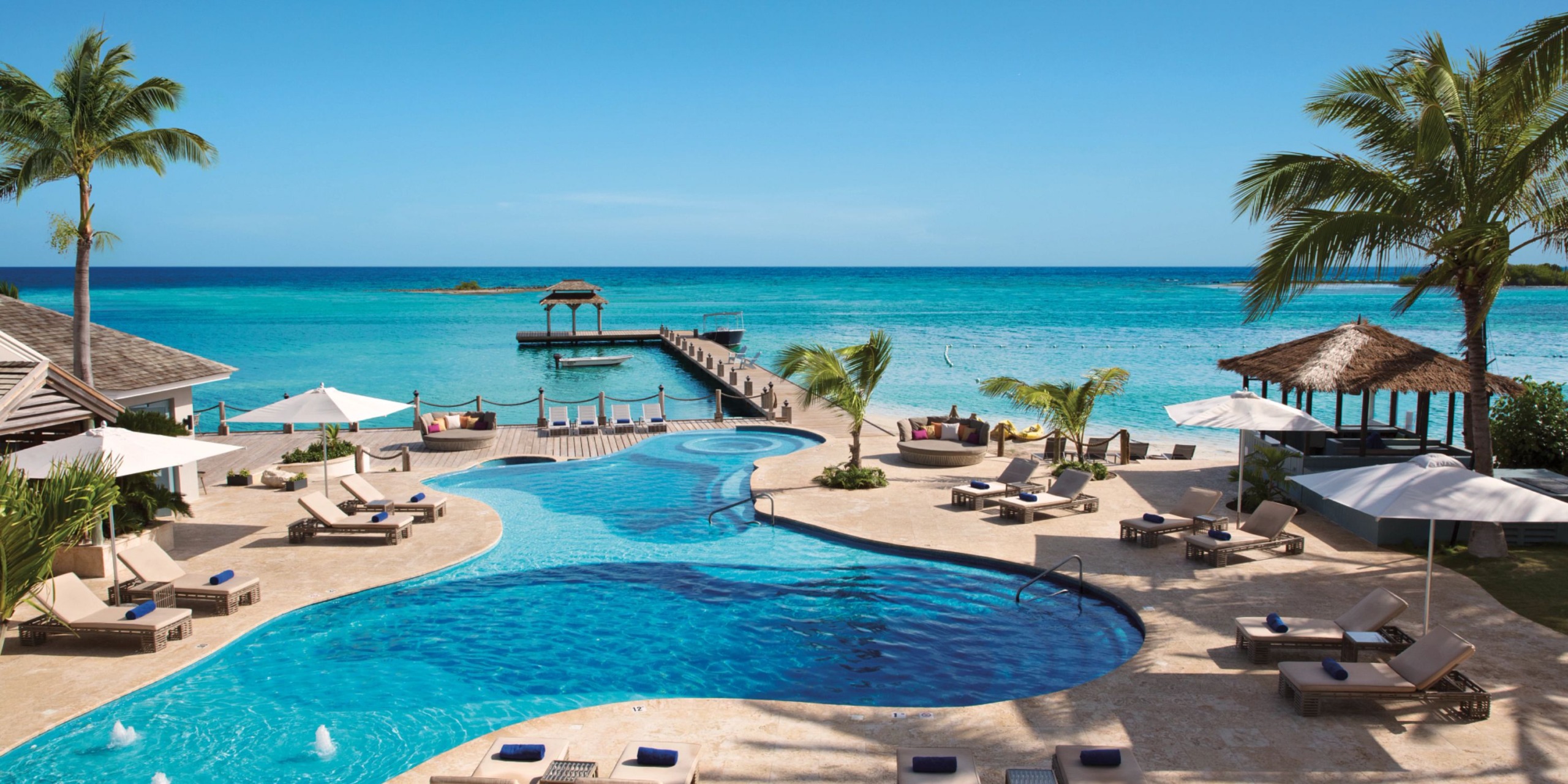 Immerse yourself in the luxury of Zoetry Montego Bay, a tropical resort featuring a spacious, winding pool encircled by comfortable lounge chairs and swaying palm trees, adjacent to a dock that stretches out into the pristine blue ocean under a radiant sky.