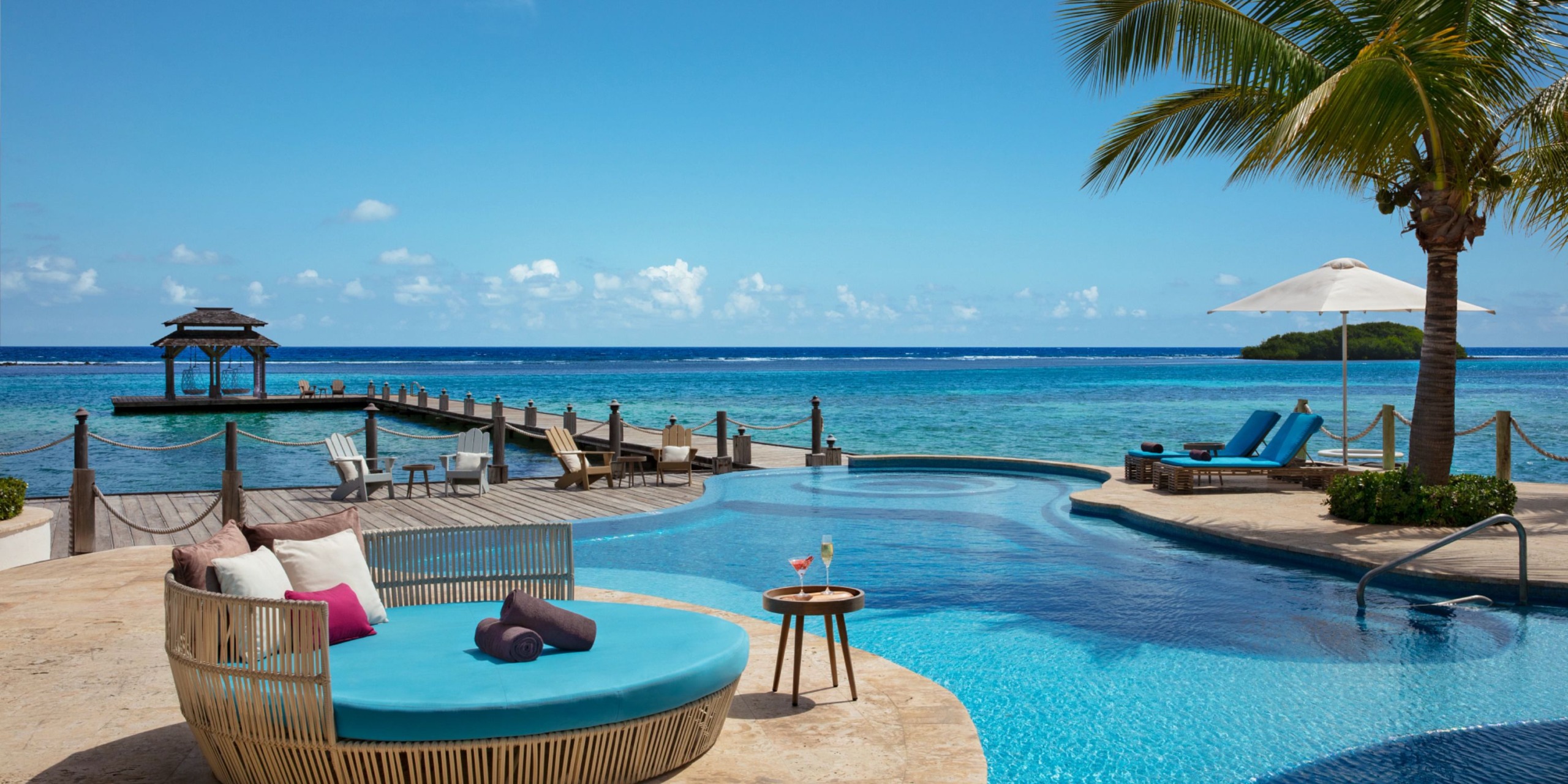 At Zoetry Montego Bay, you can relax on lounge chairs by the infinity pool, which offers breathtaking views of a blue ocean with a palm tree and a cozy gazebo in the background.