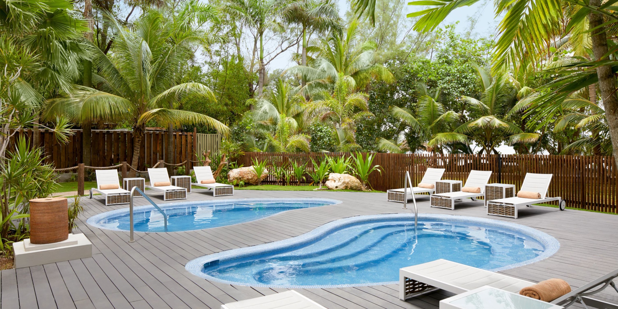 The Zoetry Montego Bay boasts an outdoor pool area featuring two kidney-shaped pools encircled by lounge chairs and lush tropical plants, with a wooden fence and trees providing a serene backdrop.